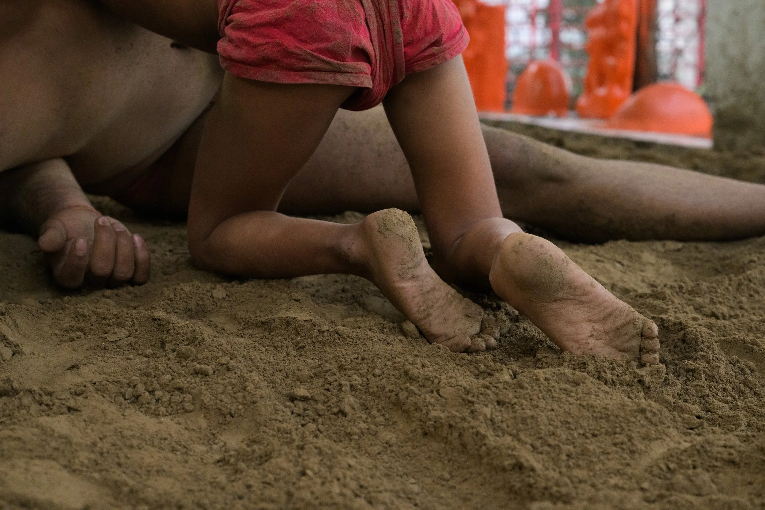 Child climbing on a sandy indoor play area, with sand on their hands and feet and an orange climbing structure in the background.