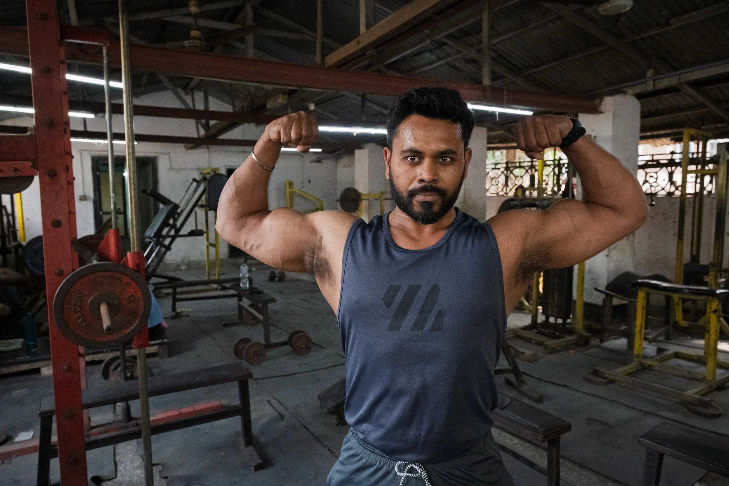 A man in a blue sleeveless shirt flexes his biceps in a gym with weightlifting equipment and benches in the background.