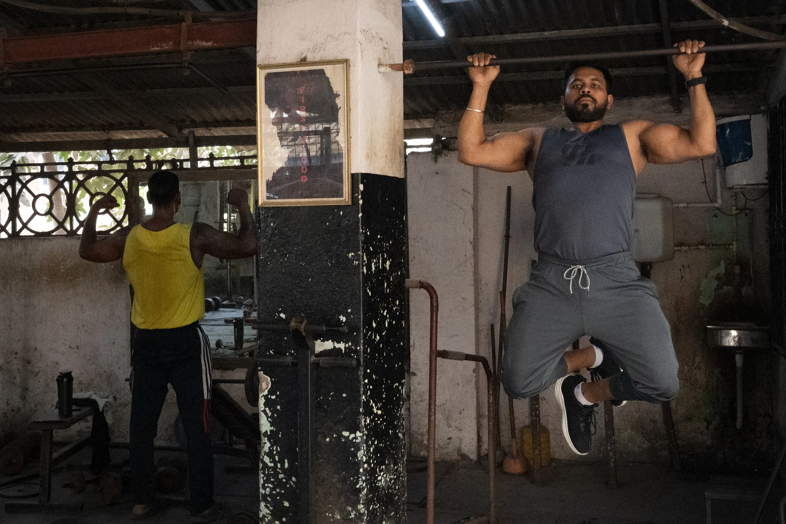 Man lifting a barbell with both arms in a gym, with another person in a yellow tank top working out in the background.