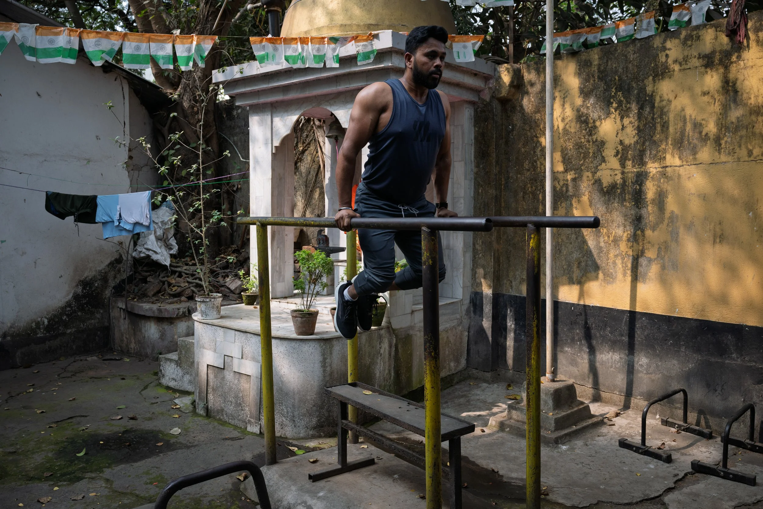 A man in a sleeveless athletic shirt and pants is doing a dip exercise on parallel bars outdoors. There are potted plants, laundry hanging, and a small shrine or temple structure in the background.