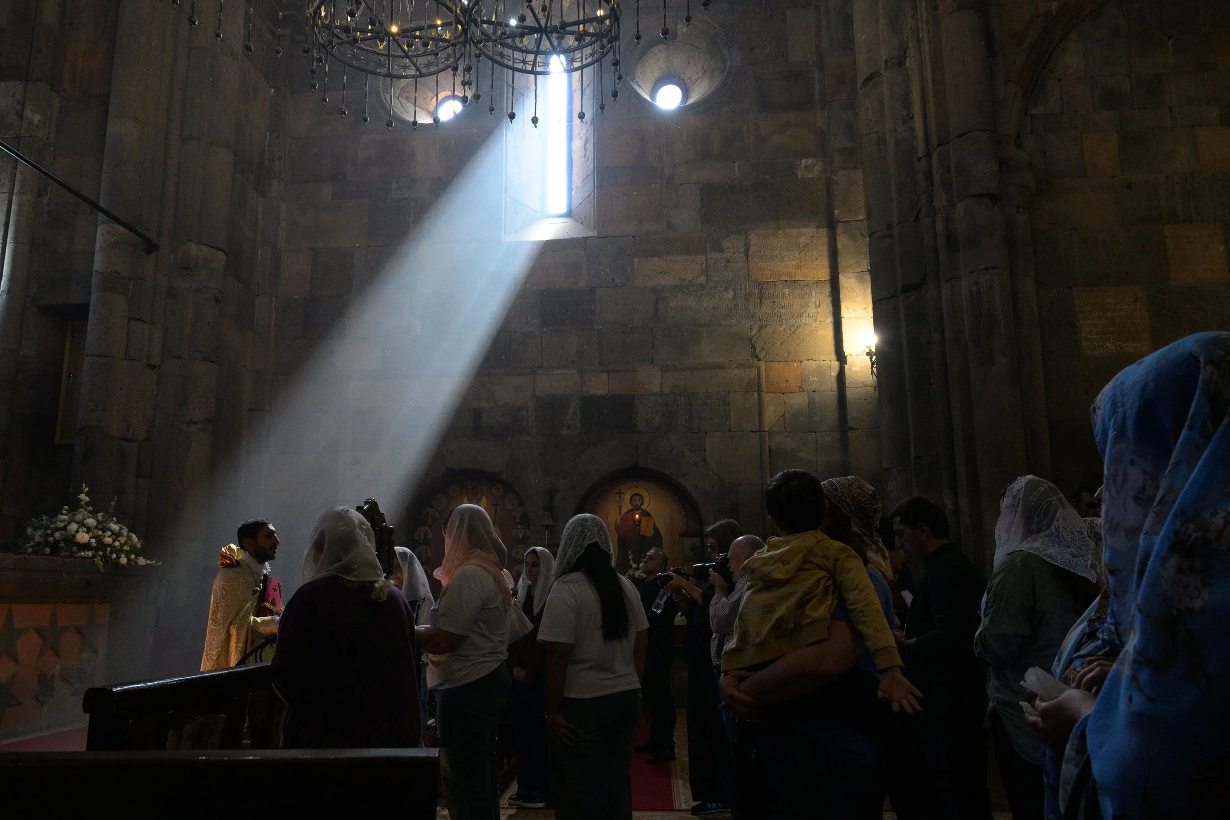 Inside a church with a large congregation. Sunlight streams through a narrow window onto the congregation, illuminating the space with a beam of light. People are gathered, some wearing head coverings, and there is religious iconography on the walls.
