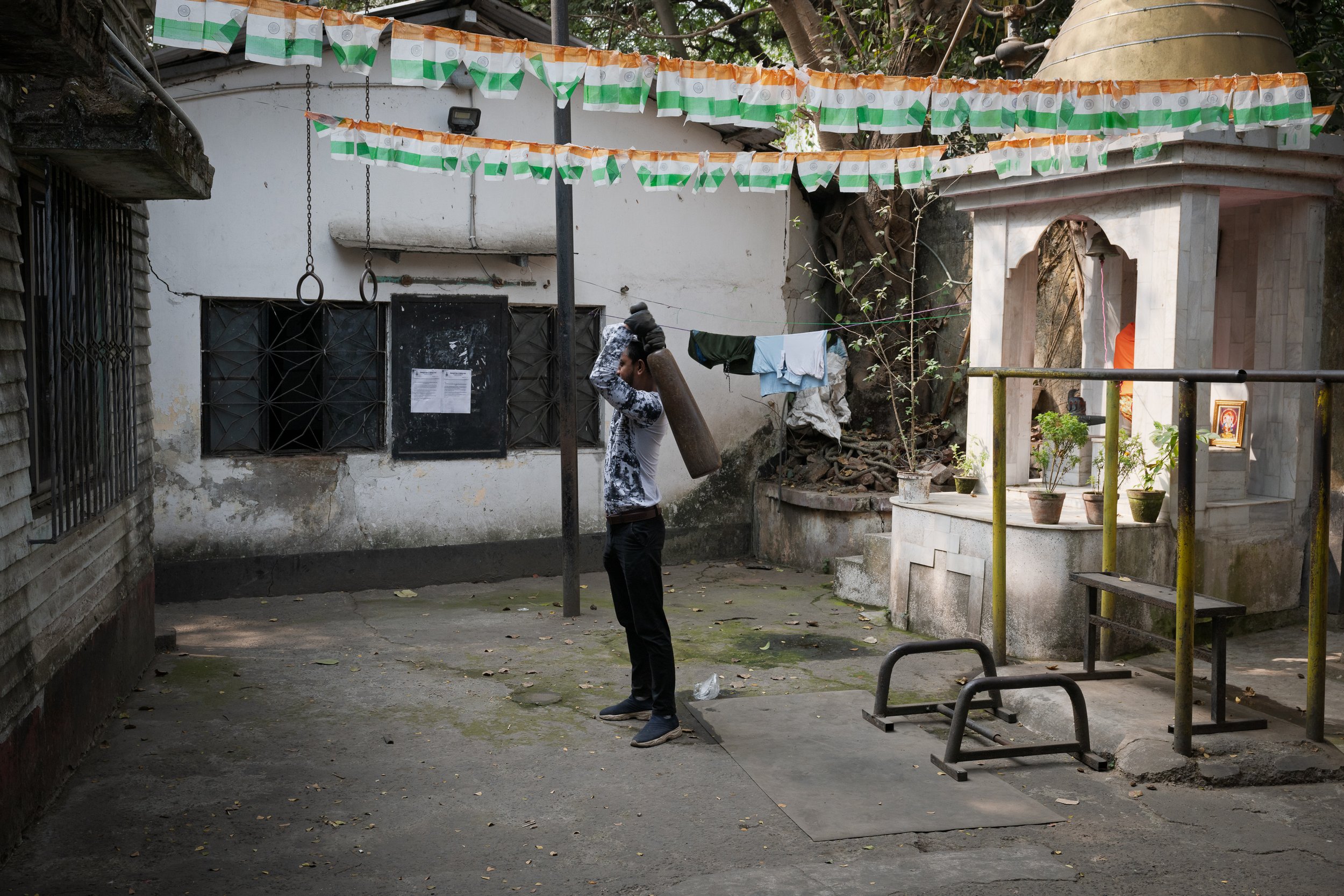 A man in dark pants and a light patterned shirt carrying a large object on his shoulder in an outdoor courtyard. The courtyard has hanging flags, a small shrine with potted plants, and laundry hanging on a clothesline. The area appears aged with some
