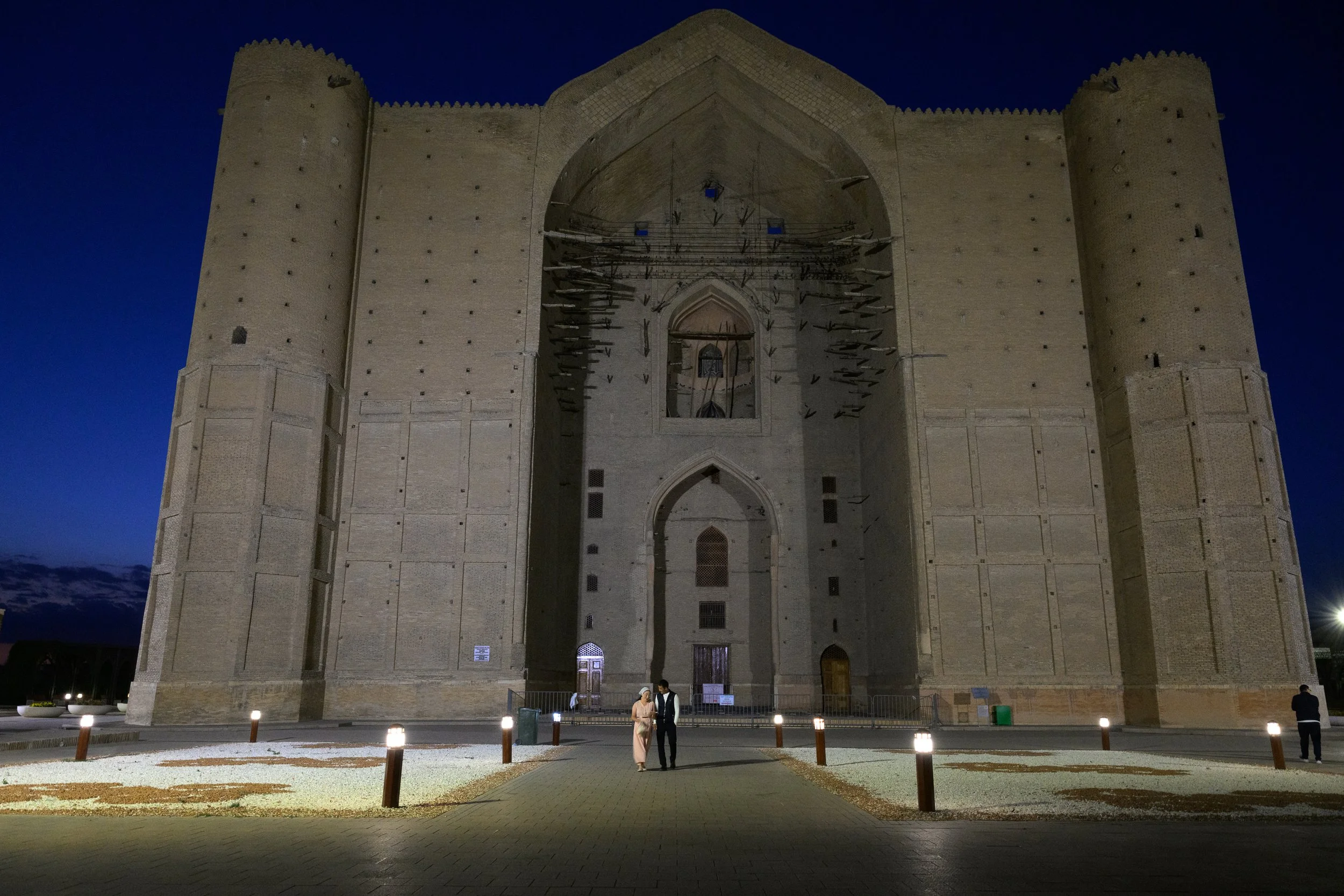 A large historic castle with two rounded towers on the sides, illuminated at night, with a pathway and a few people in front.