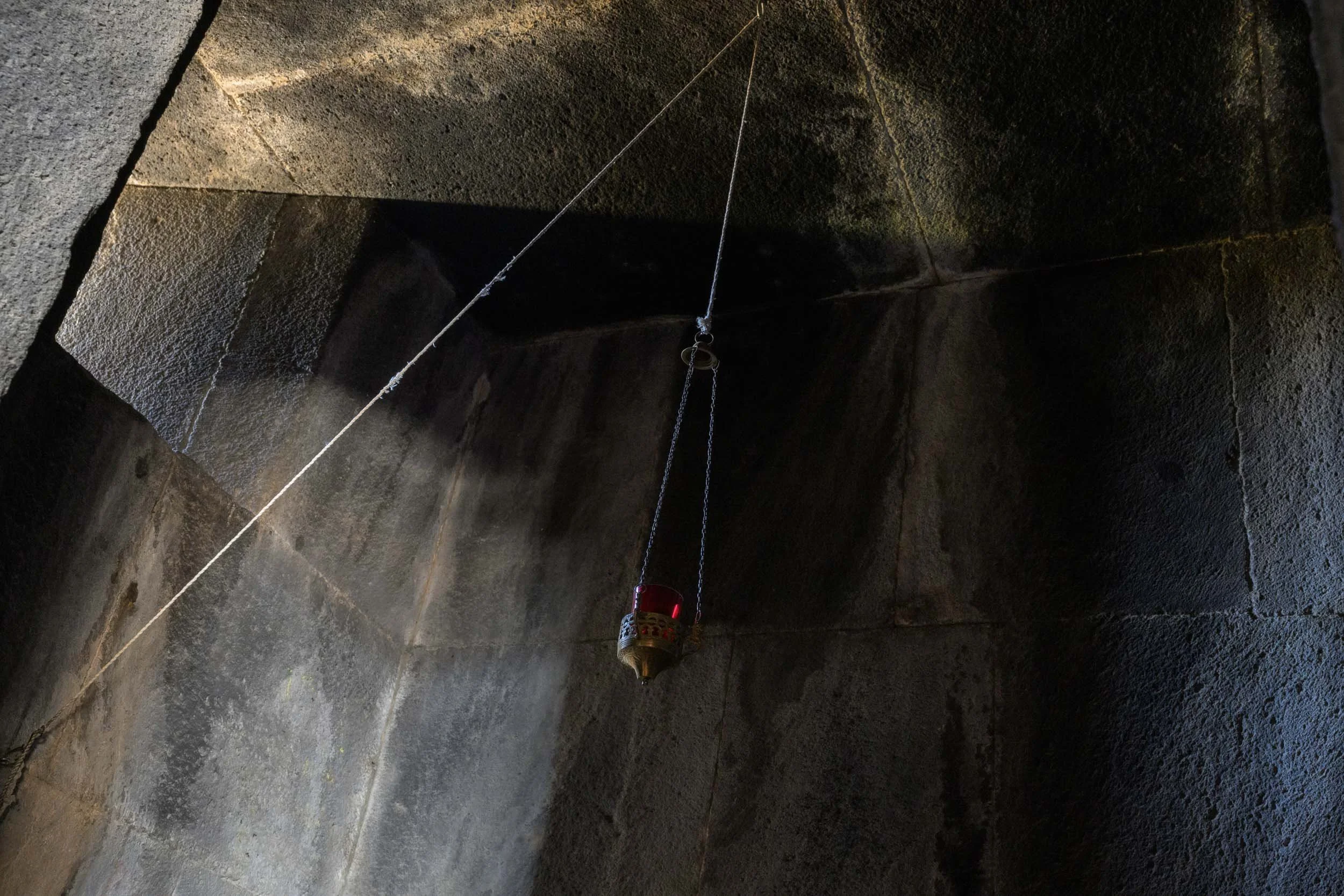 Suspended lamp illuminated by a shaft of light inside a dark ancient stone monastery interior.