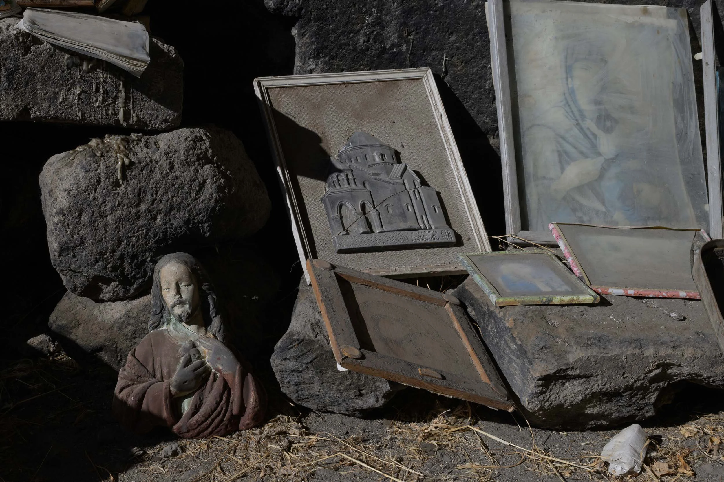 A small sculpture of Jesus Christ shows him with closed eyes and a solemn expression, wearing a robe with a sash across the chest, and holding a small bird in his right hand. The sculpture is placed among rocks and stones, with a framed picture of a 