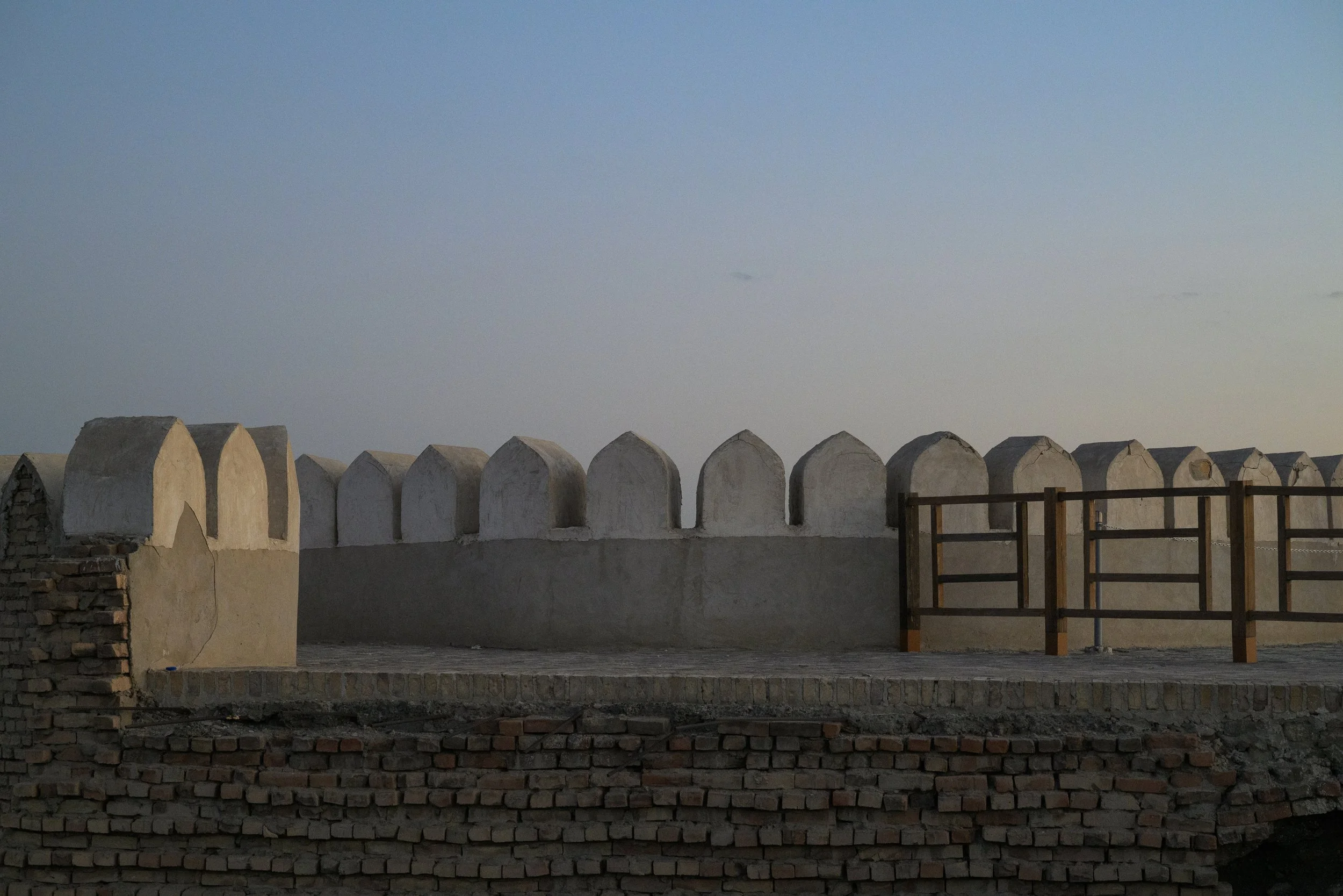 A section of a historic wall with white, domed battlements, and a wooden railing on a brick and stone base against a clear sky.