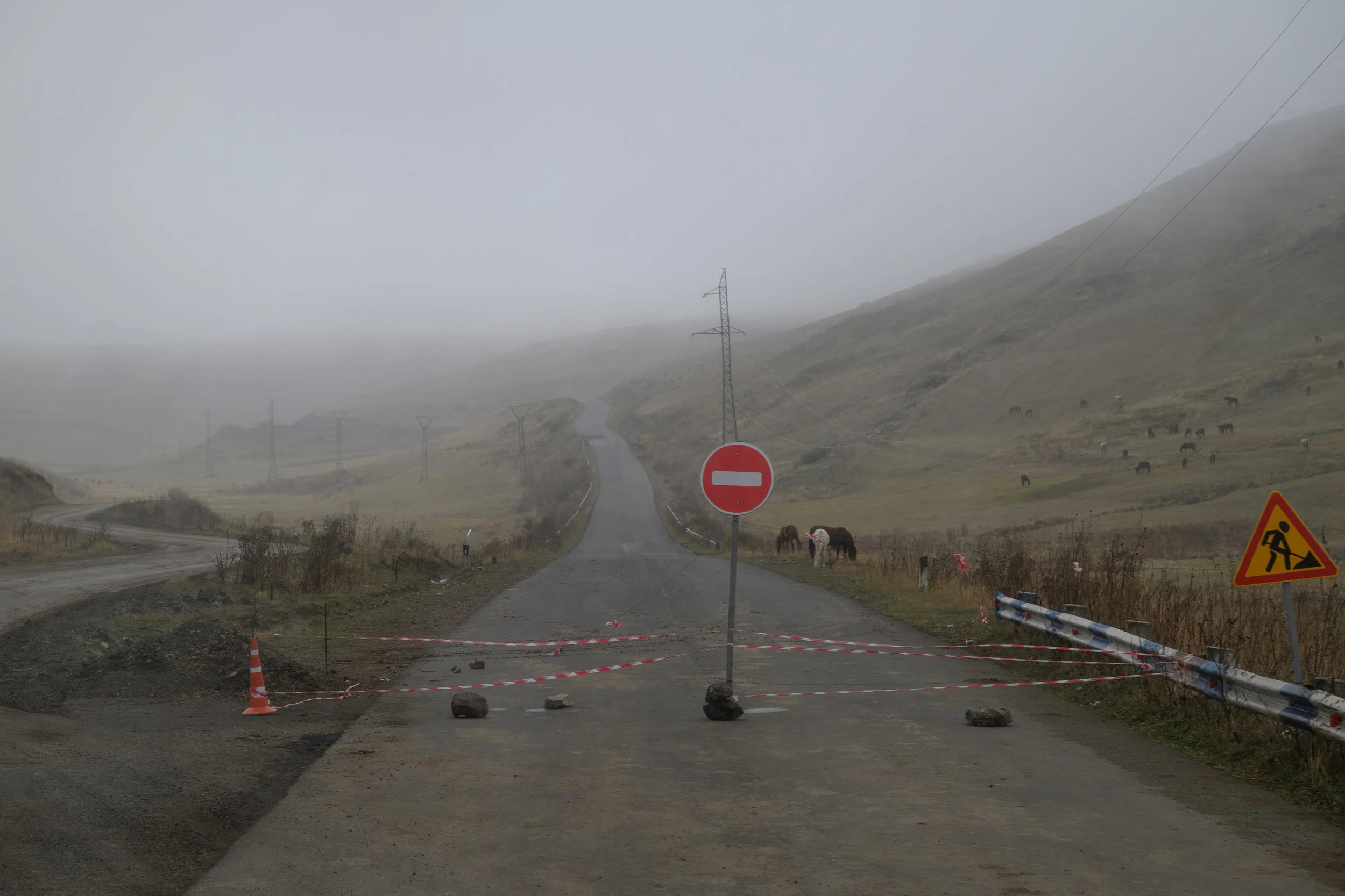 A rural mountain road blocked by rocks and red and white tape, with a stop sign and a no entry sign. There is a construction warning sign on the right side, and horses grazing on the hillside in the background. The scene is foggy and overcast.