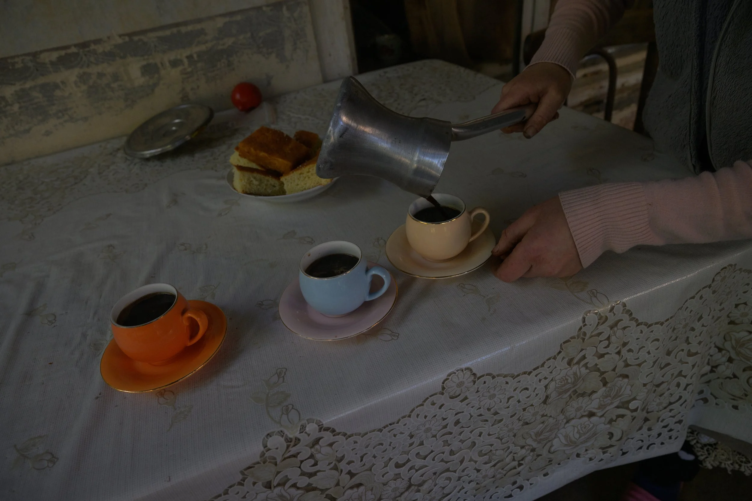 Person pouring coffee into a cup on a table with three cups of coffee, a plate with slices of cake, and a lace tablecloth.