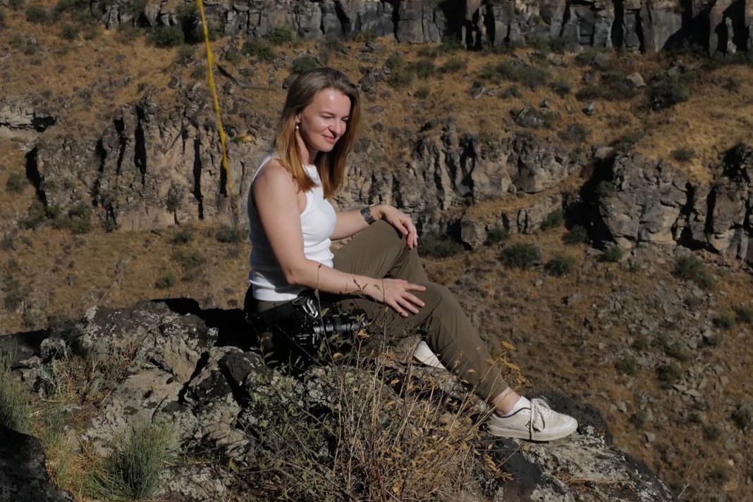 A woman sitting on a rocky ledge in a desert landscape with cliffs in the background, holding a camera.
