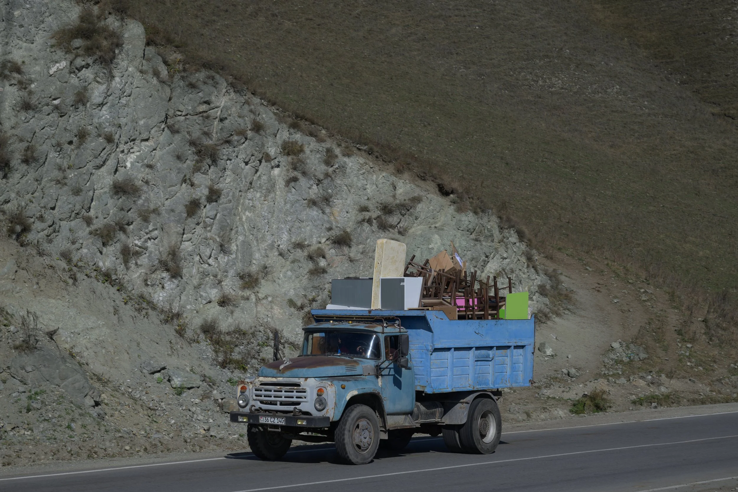 An old blue truck driving on a road with a rocky hillside in the background, loaded with furniture and chairs.
