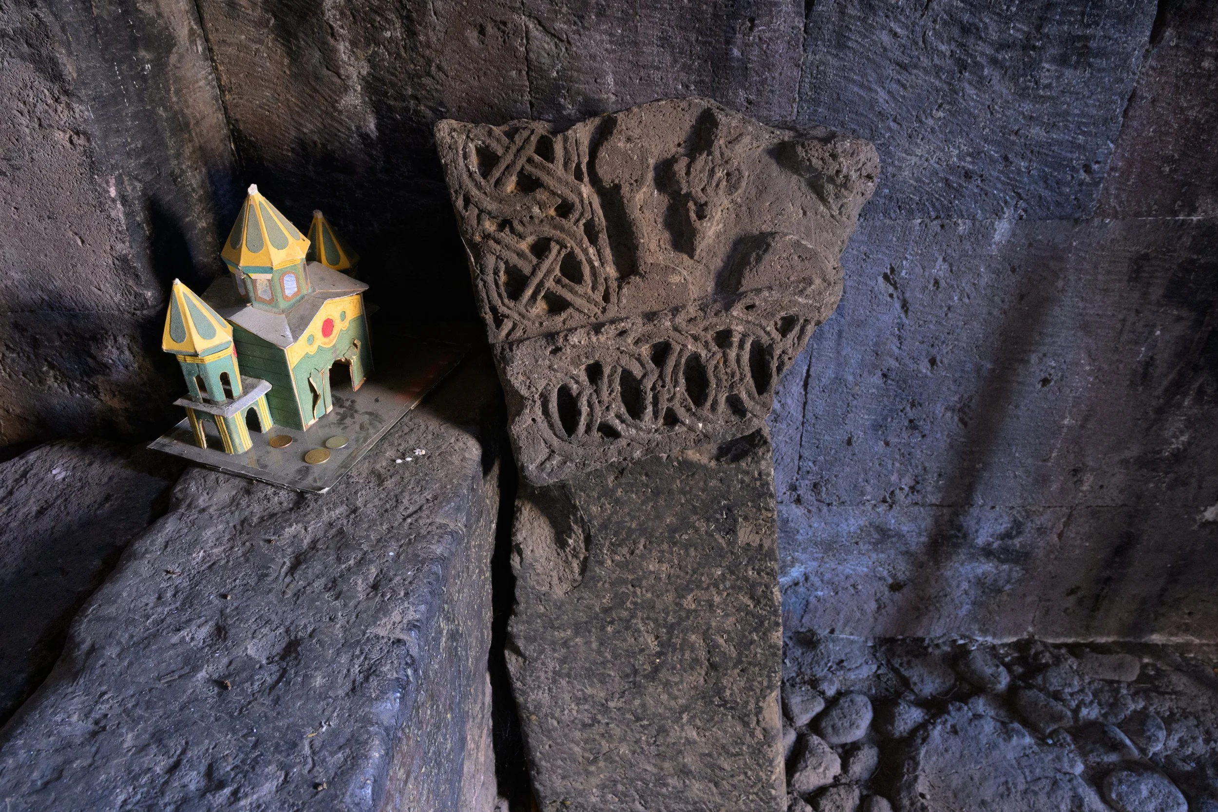 A small colorful paper model of a castle with coins on a rock, next to a carved stone cross in a dark, rocky environment.