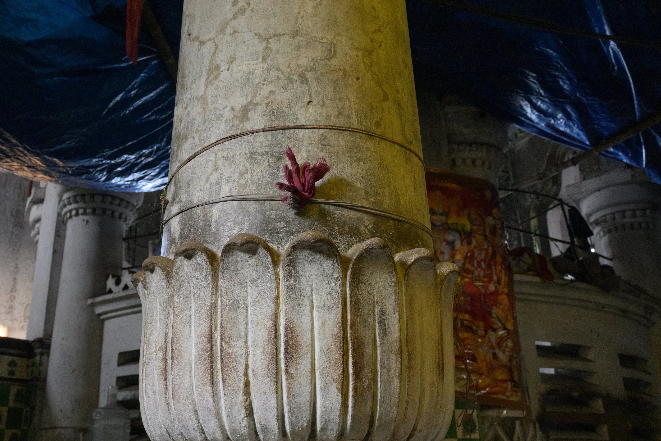 Close-up of a stone pillar inside a temple, with a pink flower and thin wire tied around it. Background shows historical carvings and dark blue drapes or tarps.