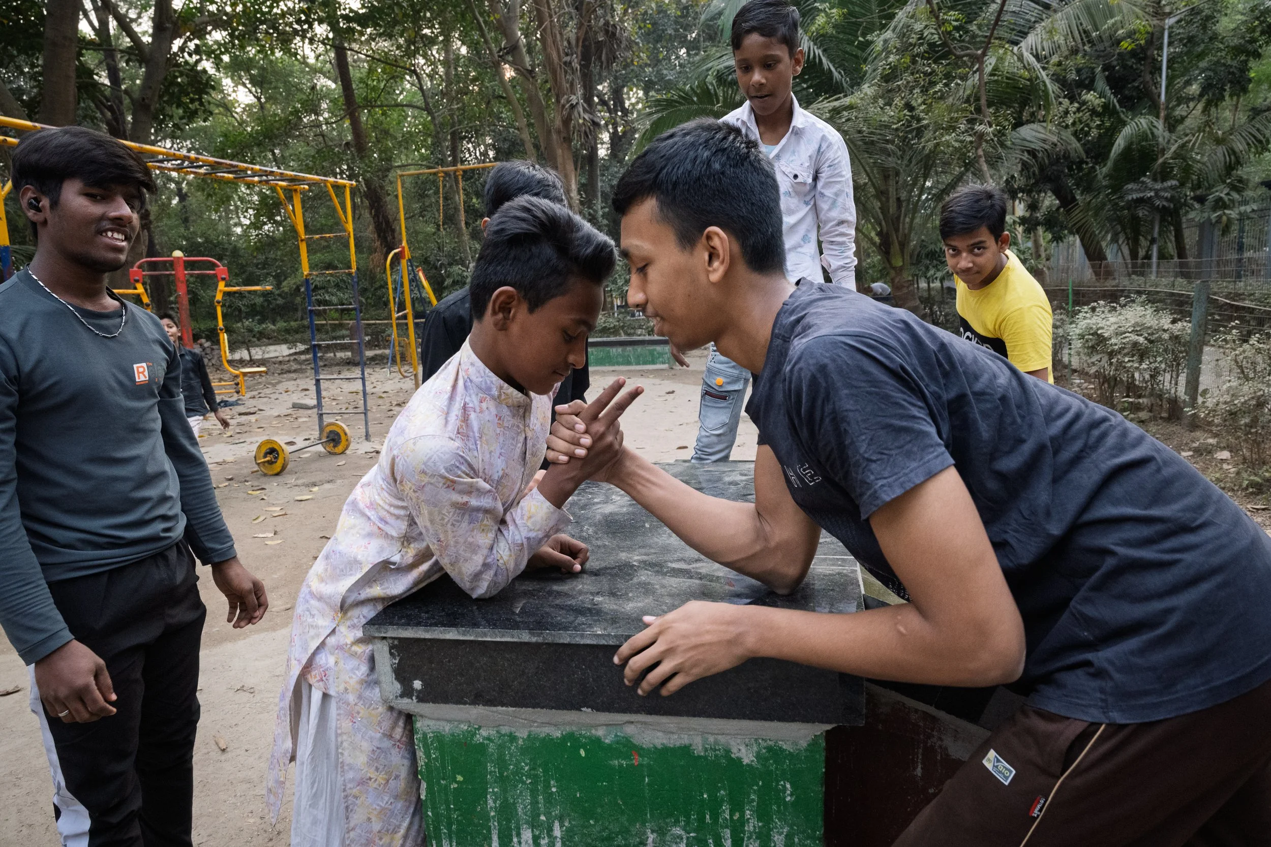 Two young men are arm wrestling at a park table, surrounded by children and teenagers watching, with trees and playground equipment in the background.