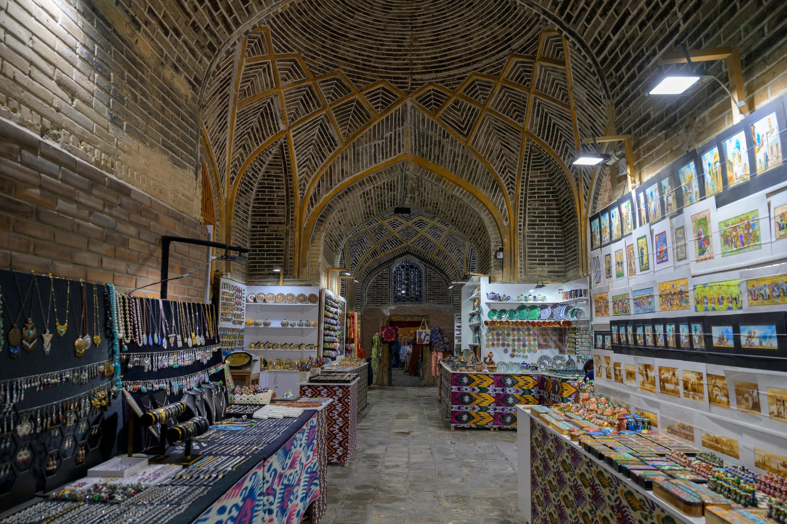 Art and craft market in a historic indoor building with arched ceiling, displaying jewelry, paintings, and decorative items on tables and walls.