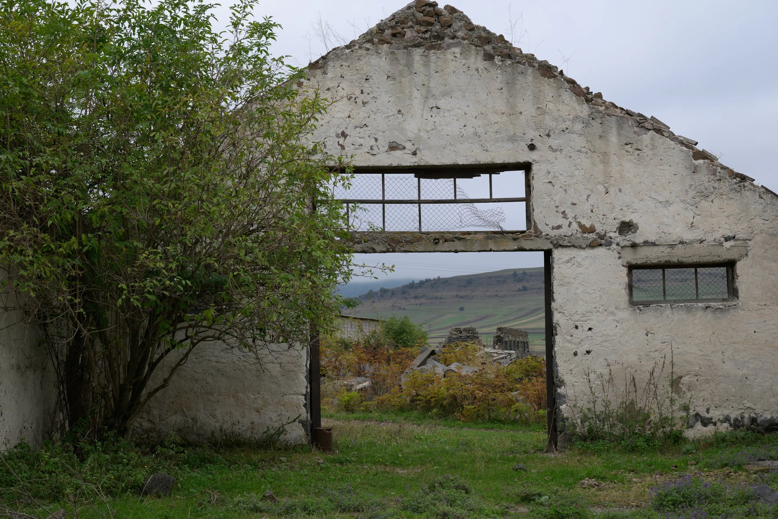 An old, abandoned brick building with large rectangular openings and a smaller window, surrounded by overgrown vegetation and a rural landscape in the distance.