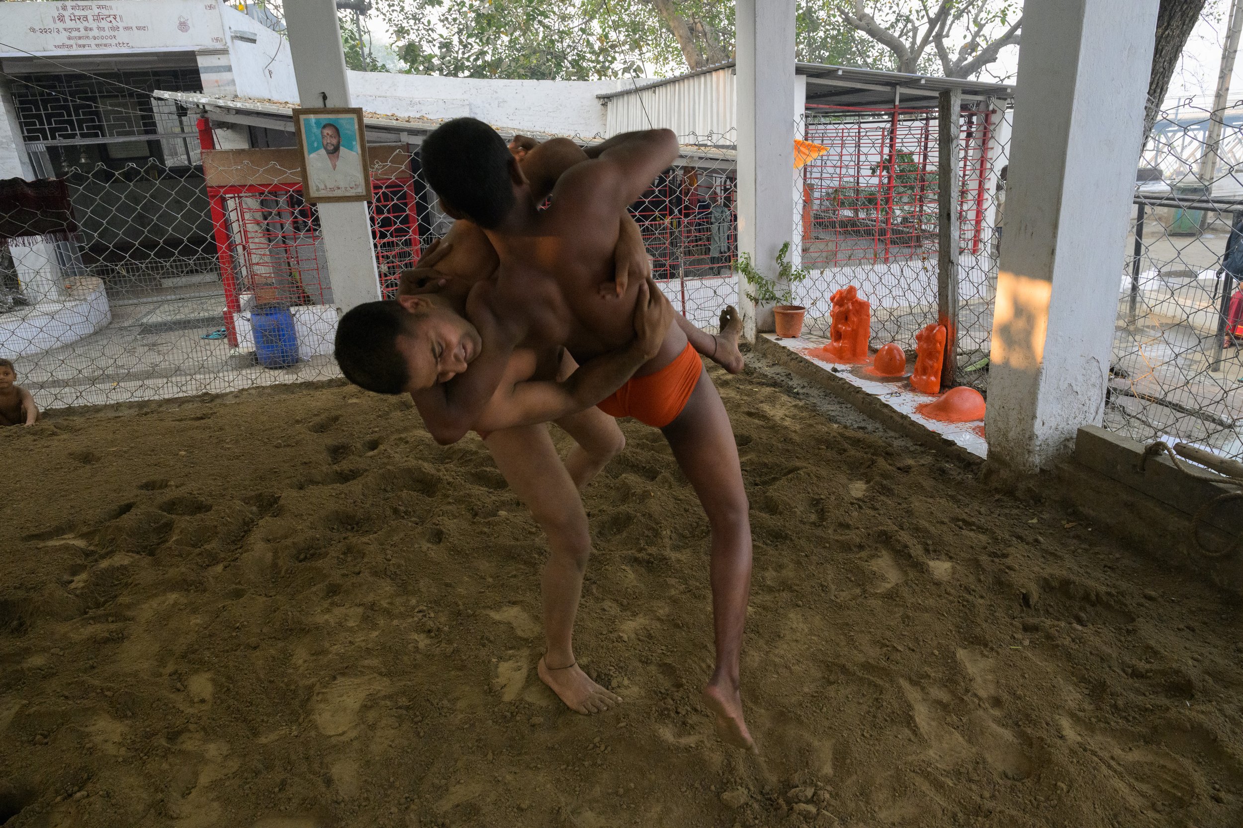 Two young boys practicing wrestling in a dirt or sand training area, with a background of a wire fence, potted plants, and statues. One boy has lifted the other in a wrestling move.
