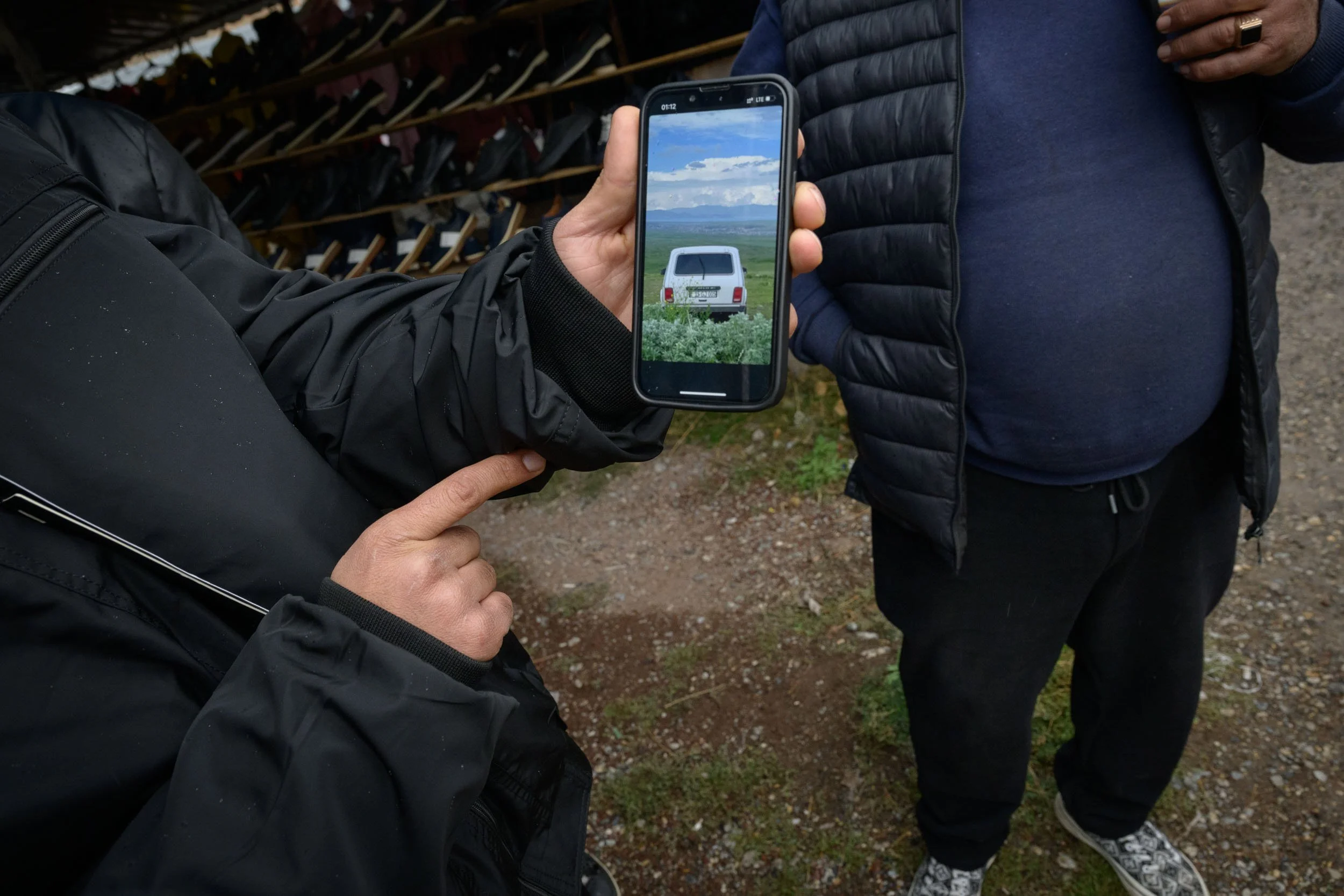 A person is holding a smartphone taking a picture of an old white van with a rounded back and tail lights in a green field under a cloudy sky. Another person is standing nearby, wearing a black puffer vest and blue sweatshirt. In the background, ther