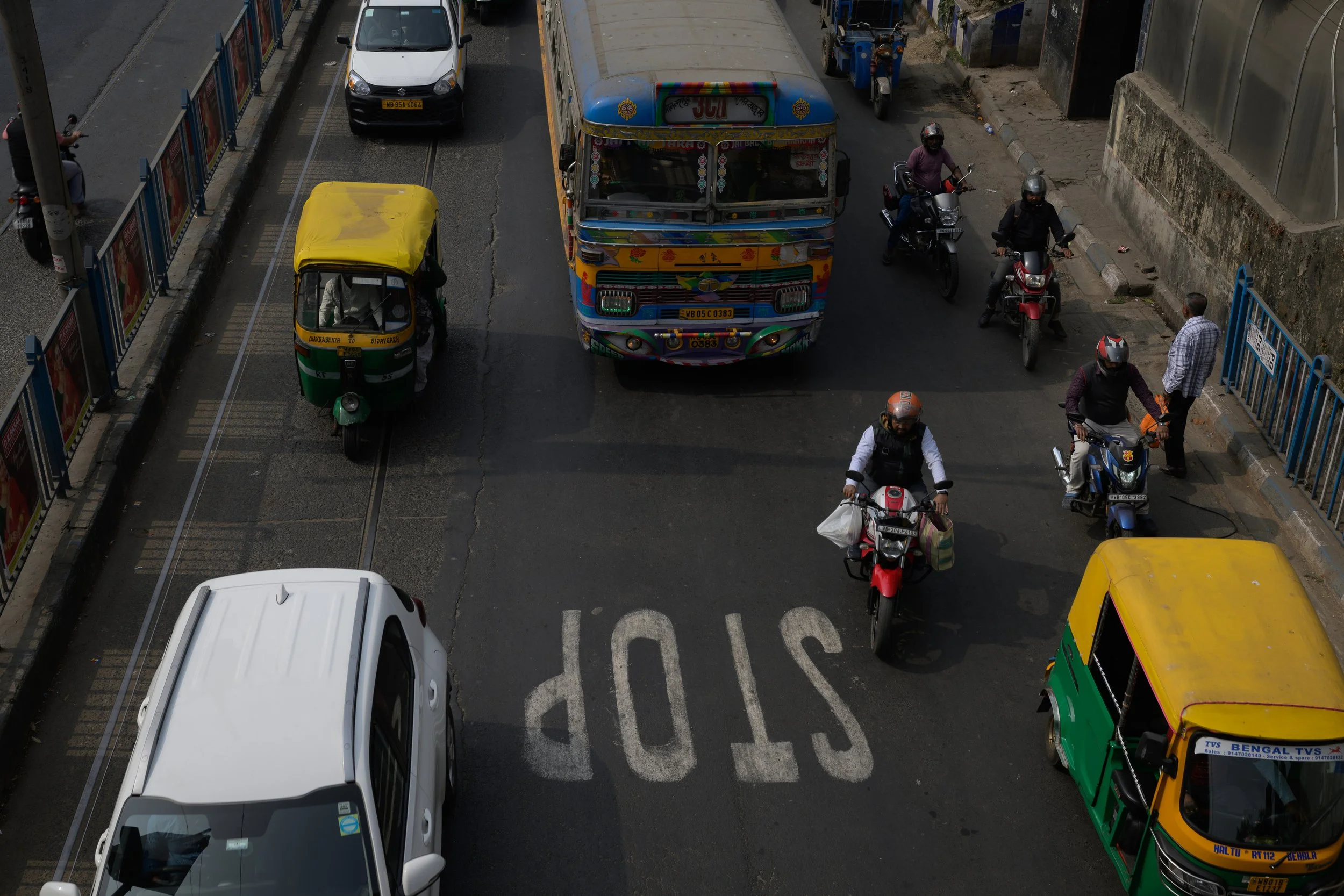 A busy street with various vehicles, including cars, auto rickshaws, and motorcycles, seen from above. A large, colorful bus is in the center, and there's a stop sign painted on the road.