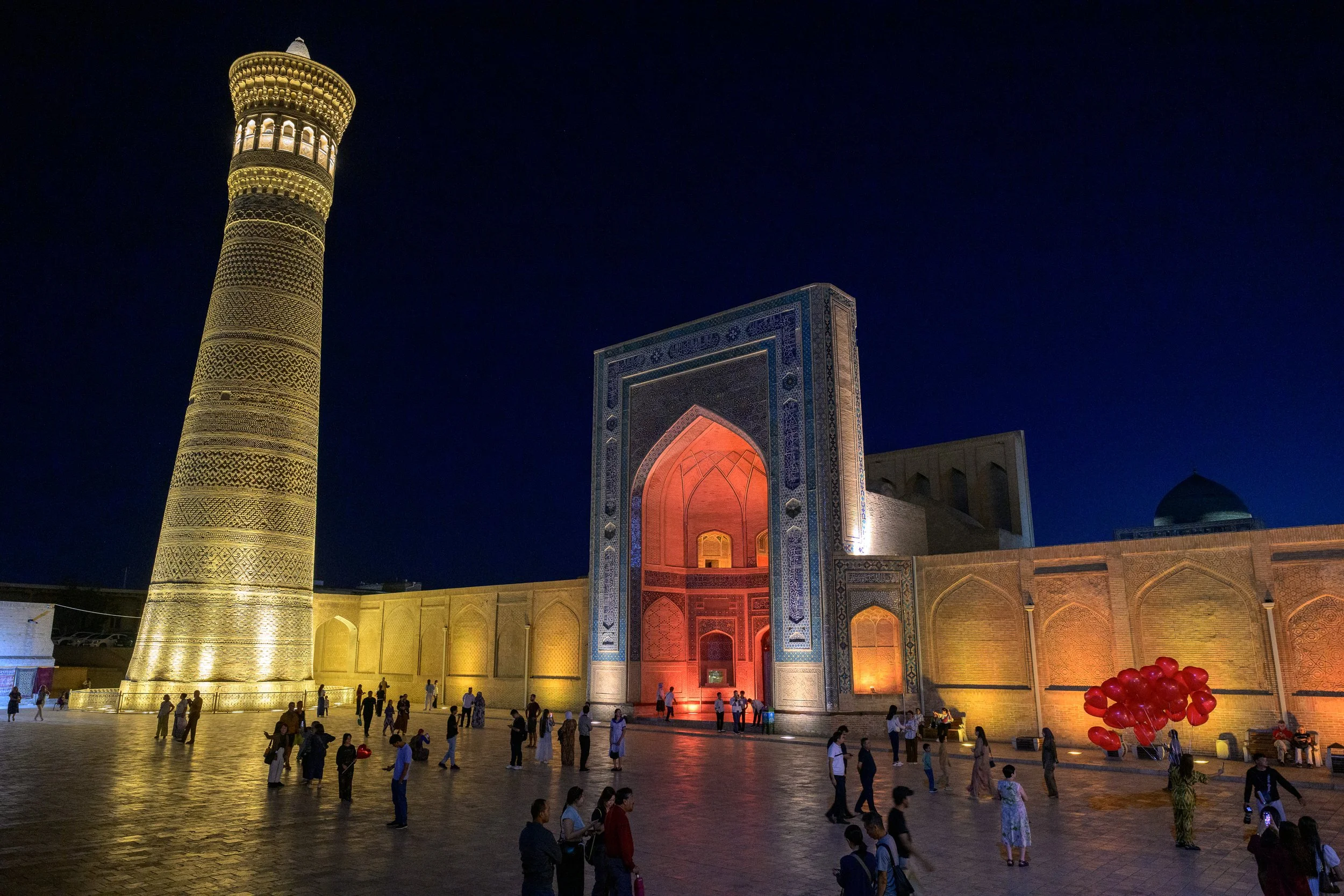 Night view of a grand Islamic architectural site with a tall illuminated minaret, an ornately decorated entrance, and people walking around the courtyard. Red balloons are visible on the right.