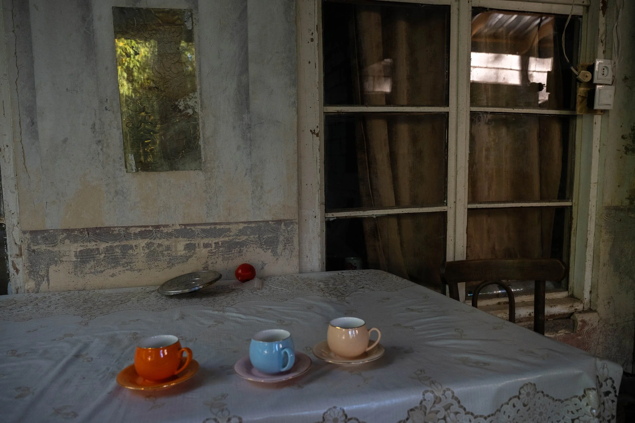 A dining table with a lace tablecloth, set with four cups and saucers in orange, blue, and white, in front of a wall with peeling paint and an old large mirror. Behind the table is a wooden cabinet with glass doors and an electrical outlet on the wall.