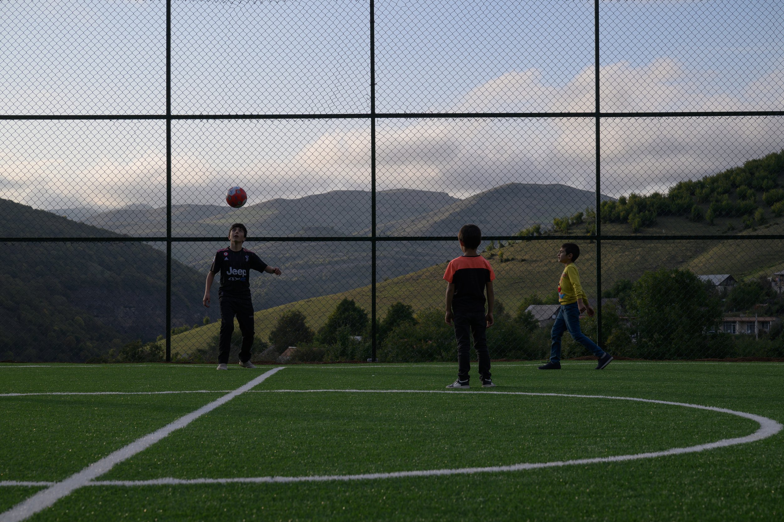 Three children playing soccer on an outdoor field with mountains in the background. One child is heading the ball, while the other two stand nearby.