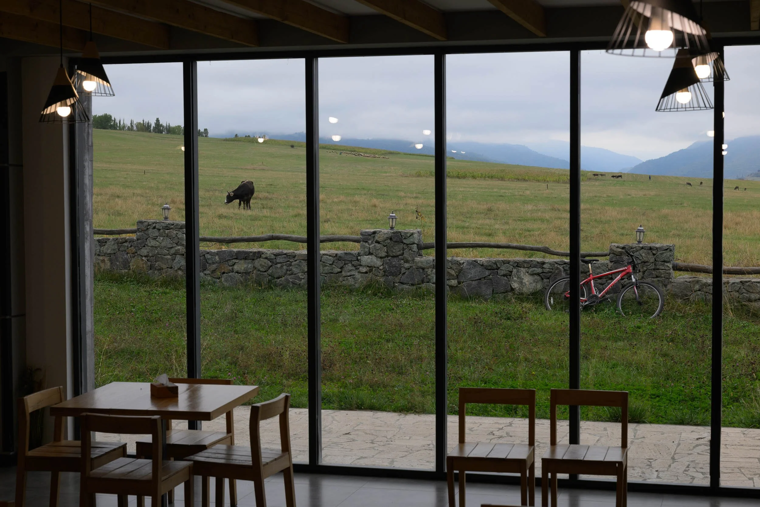 Interior view looking out a large glass window at a green field with a cow grazing, stone fence, and distant mountains, with a wooden dining table and chairs inside.