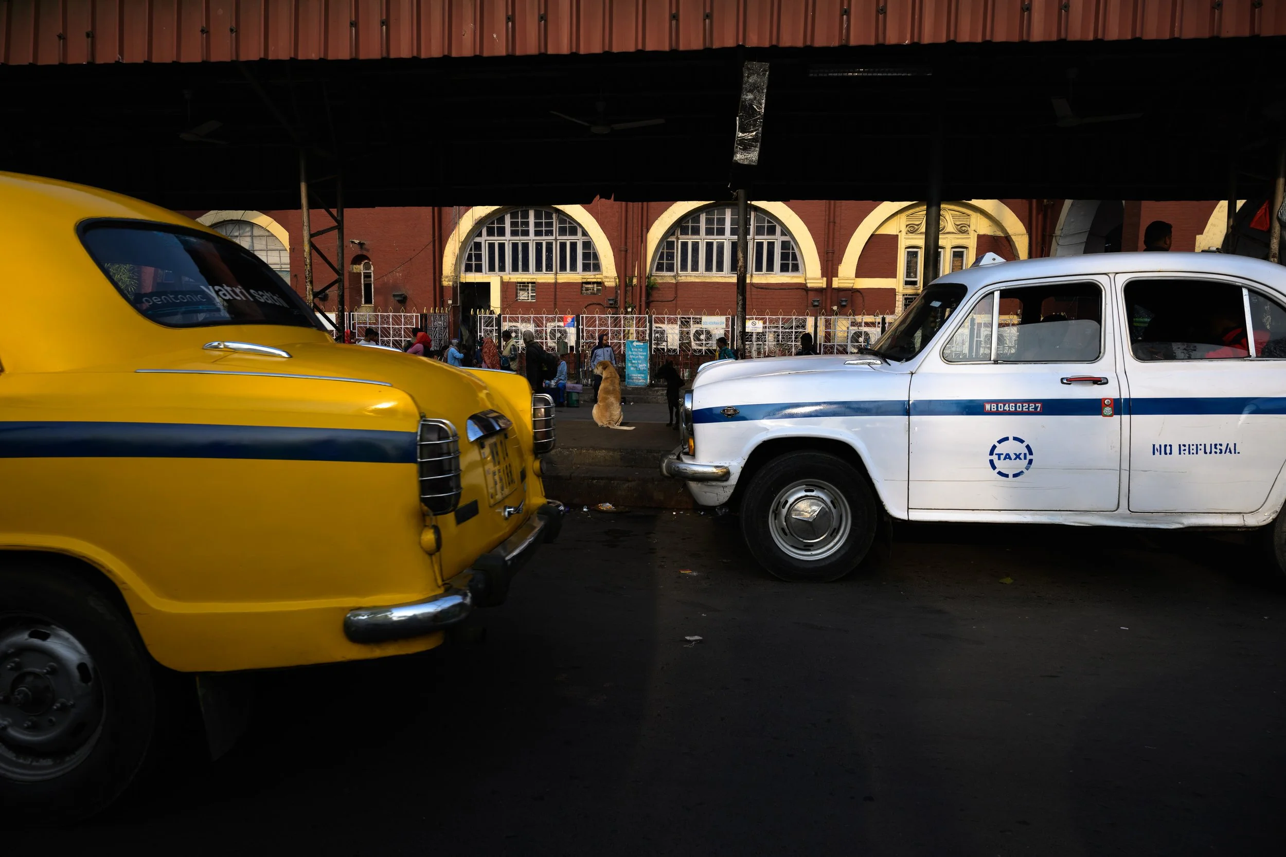 Two vintage taxis, one yellow and one white, parked under a shelter with a red building and people in the background.