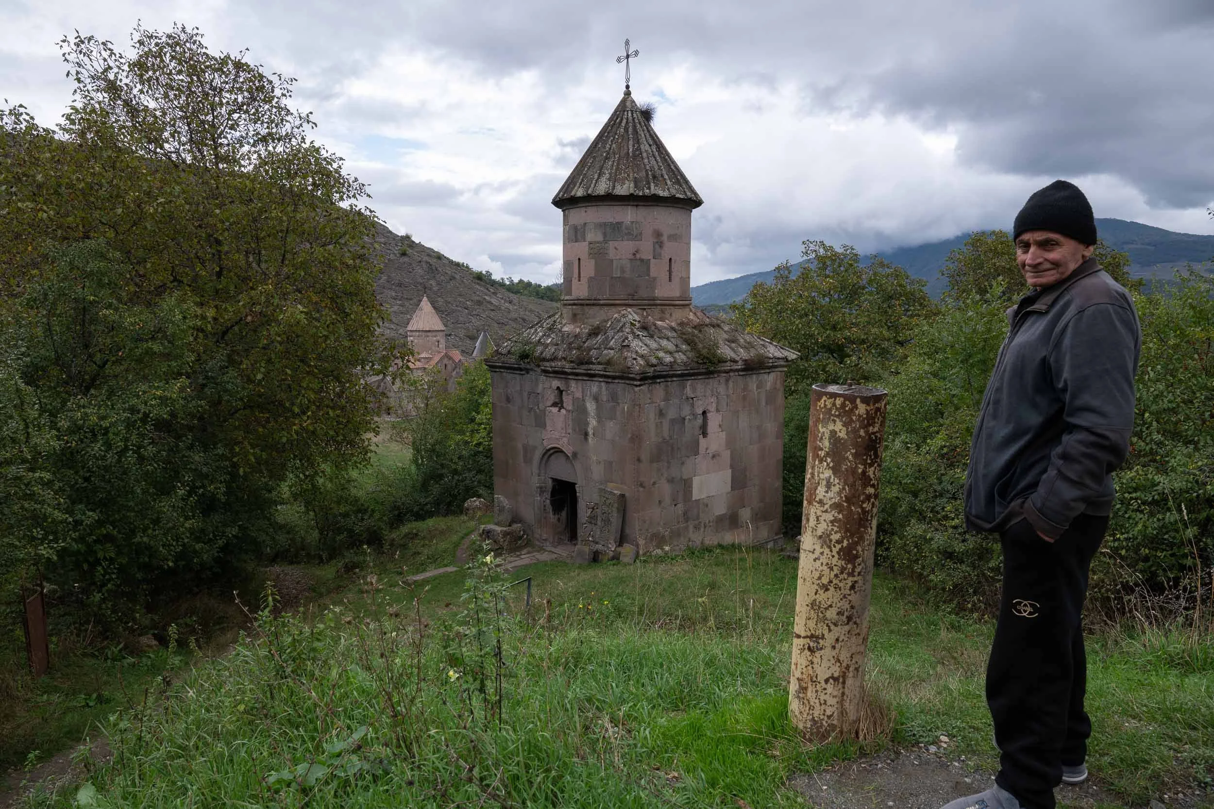 A man in a black beanie and dark jacket standing outdoors next to a small, old stone church with a conical roof and a cross on top, surrounded by trees and mountains.