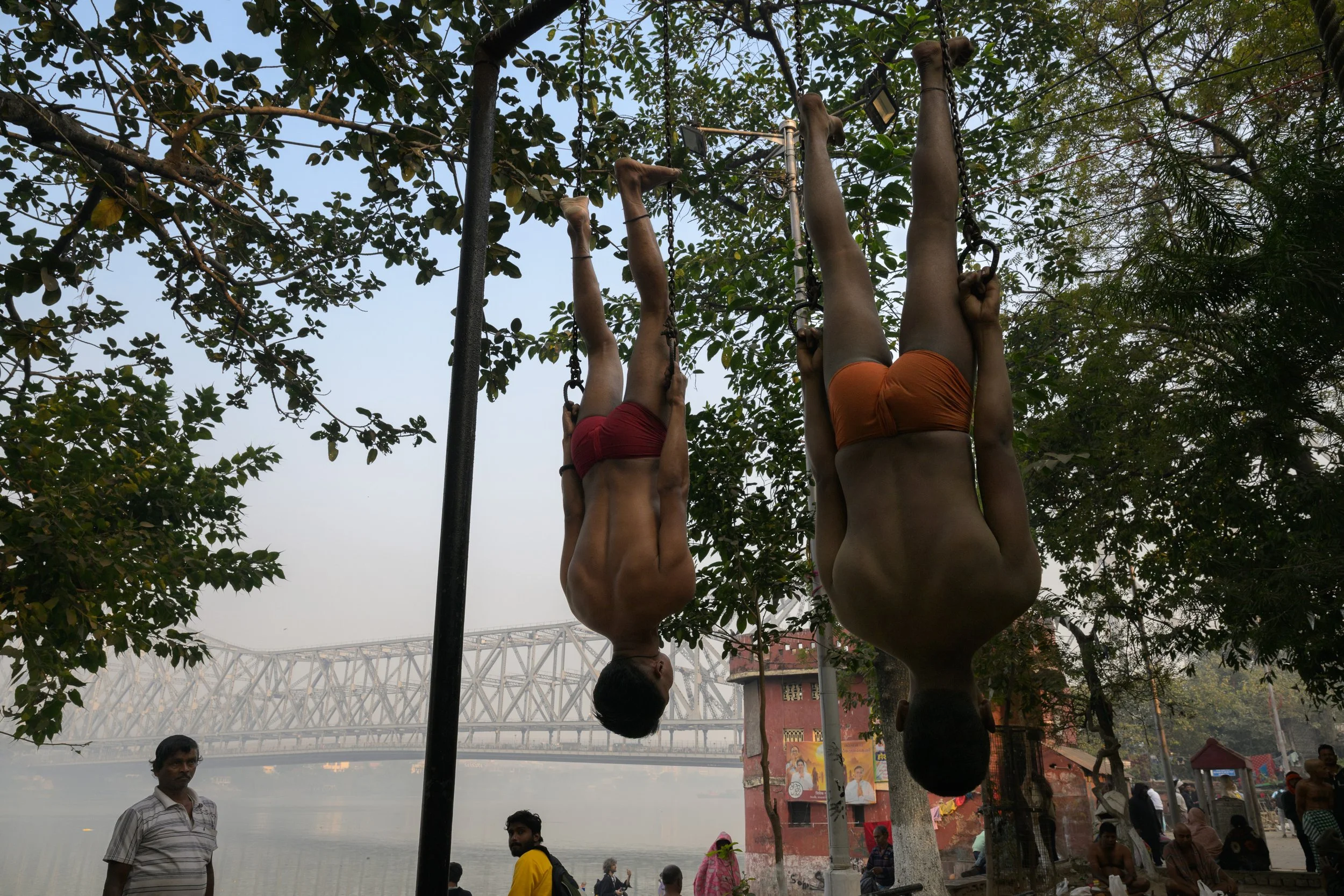 Two men hanging upside down from a gymnastic bar outdoors near a river, with trees and a bridge in the background. People are sitting and standing nearby.