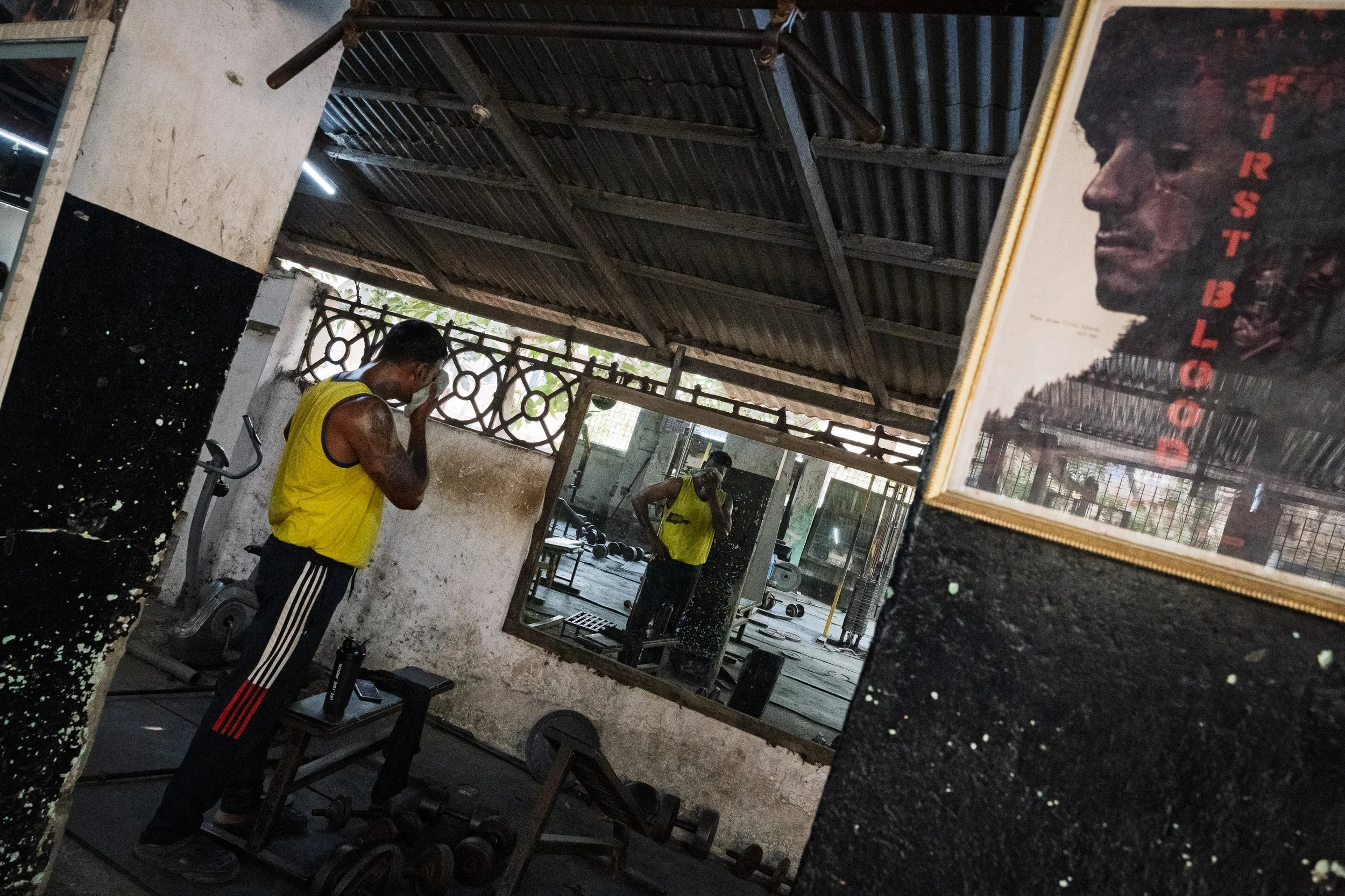 A man wearing a yellow sleeveless shirt and black track pants with red and white stripes, standing in a gym, is seen in a mirror and appears to be wiping sweat from his face. The gym has a weathered wall, various exercise equipment, and a framed post