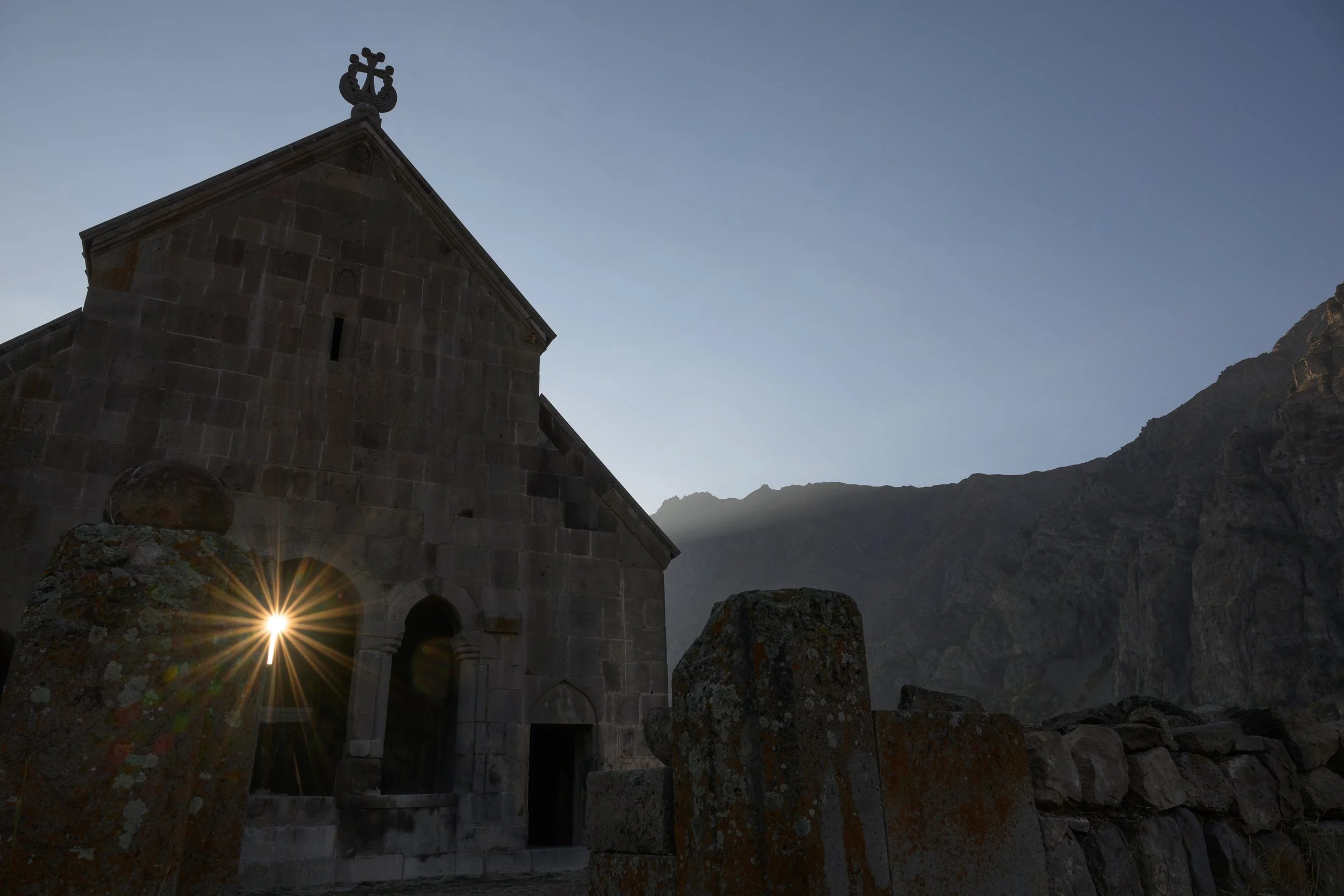 A stone church with a cross on top, with sunlight shining through an arched doorway, set against a mountainous landscape at dusk or dawn.