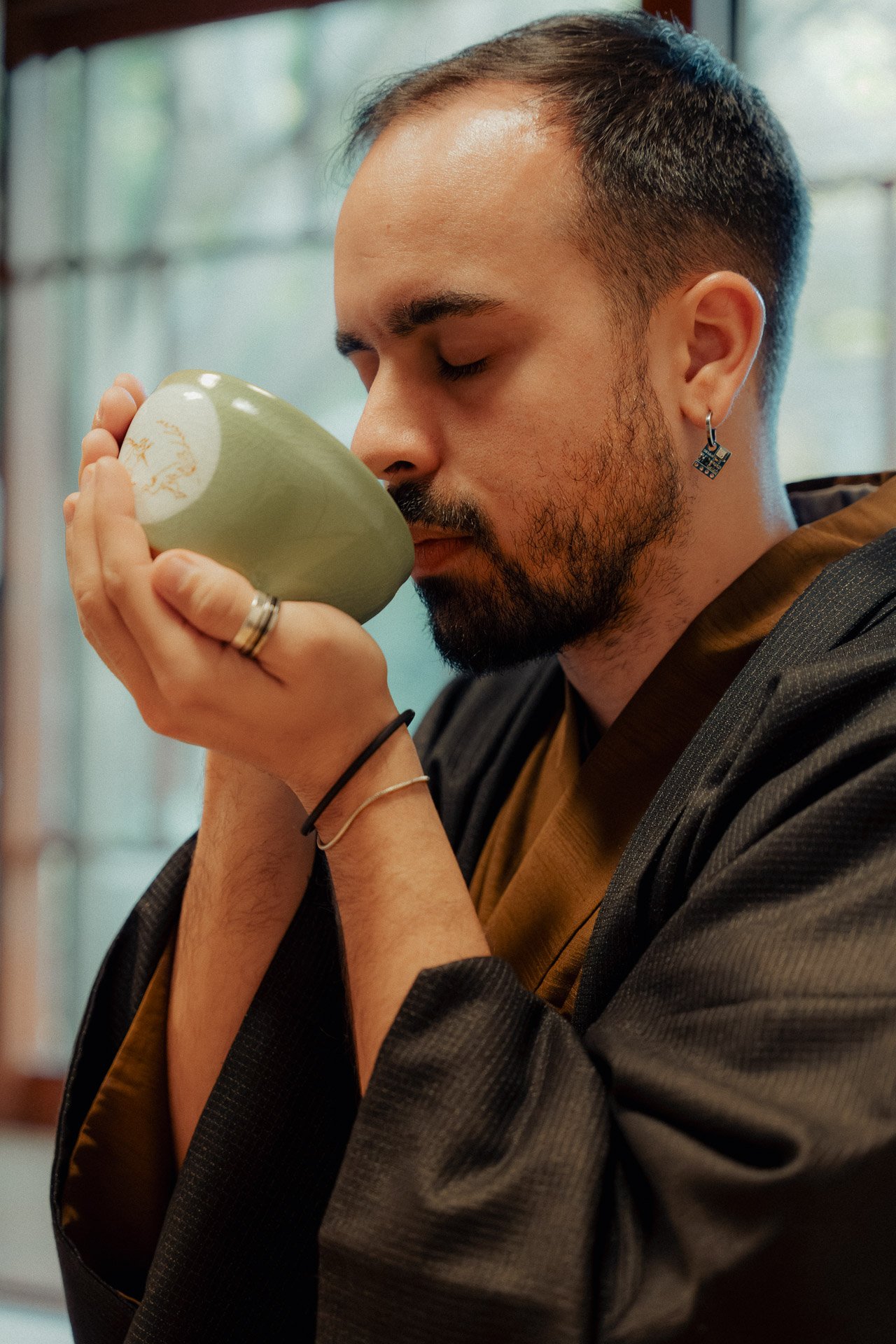 A man with a beard, earrings, and rings on his fingers is holding a green teacup close to his nose with his eyes closed, appearing to enjoy the aroma of the tea.