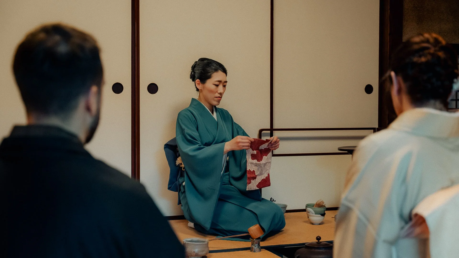 A woman dressed in traditional Japanese kimono performing a tea ceremony for two men in a tatami room.