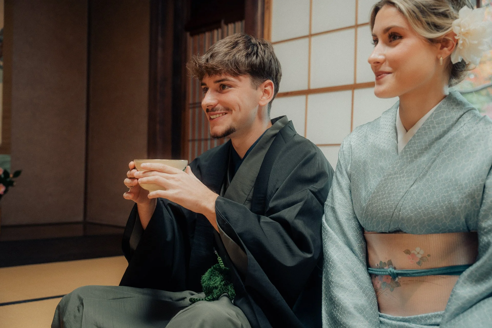 A man and a woman dressed in traditional Japanese clothing, sitting indoors, with the man holding a cup and smiling.