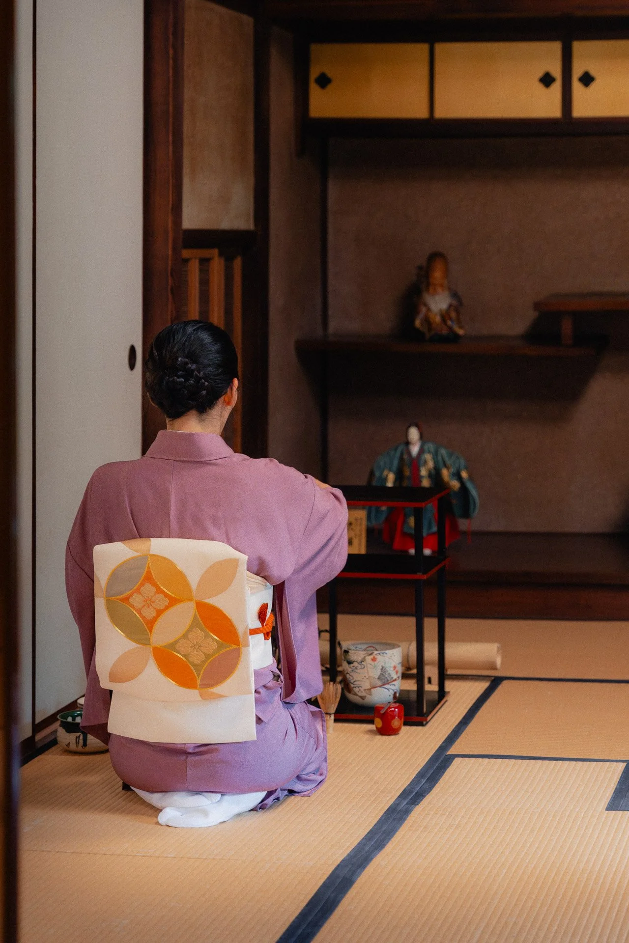 A woman wearing a pink kimono with a decorative obi kneels on tatami mats in front of a traditional Japanese altar with figures and offerings during a ceremony.