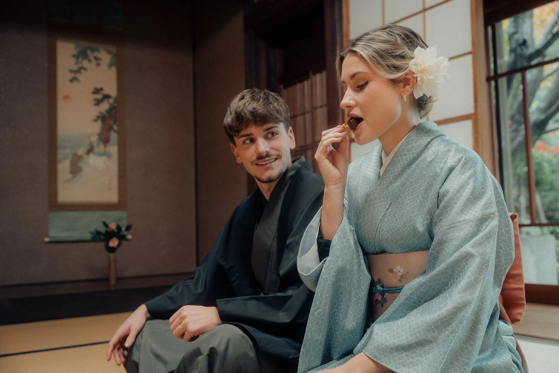 A man and woman in traditional Japanese clothing sitting in a room with tatami mats and shoji doors. The woman is wearing a kimono with a flower hair accessory and is holding a piece of candy to her mouth, looking at the man who is smiling at her.