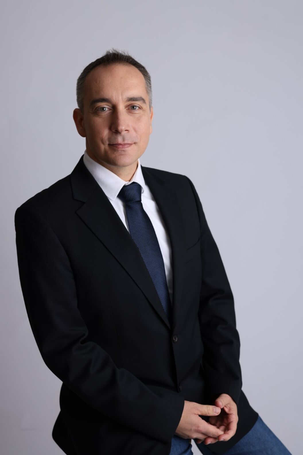 A man - Peter Olvecky, Chief Information Security Officer in a black suit, white shirt, and navy tie sitting against a plain grey background.