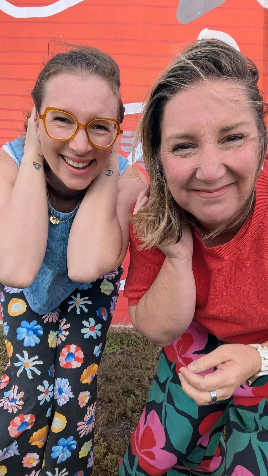 Two women taking a selfie in front of a red wall with white patterns, both smiling and leaning close to each other. One is wearing glasses, a sleeveless top, and floral pants, and the other is wearing a red shirt and floral pants.