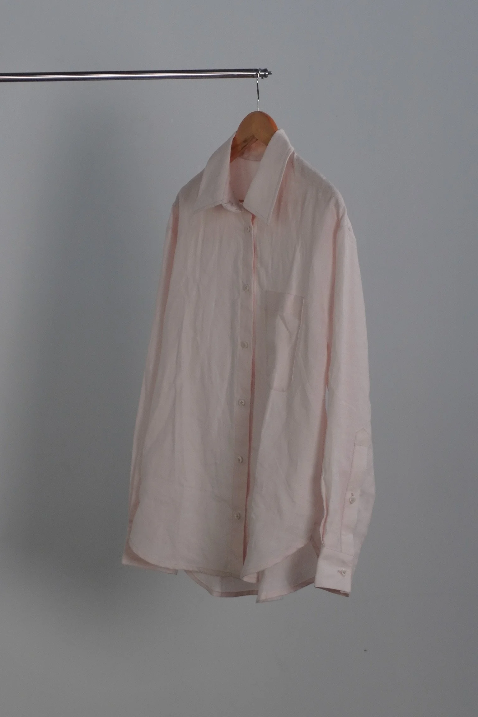 A light pink, button-up shirt hanging on a wooden hanger from a metal rod against a gray wall.