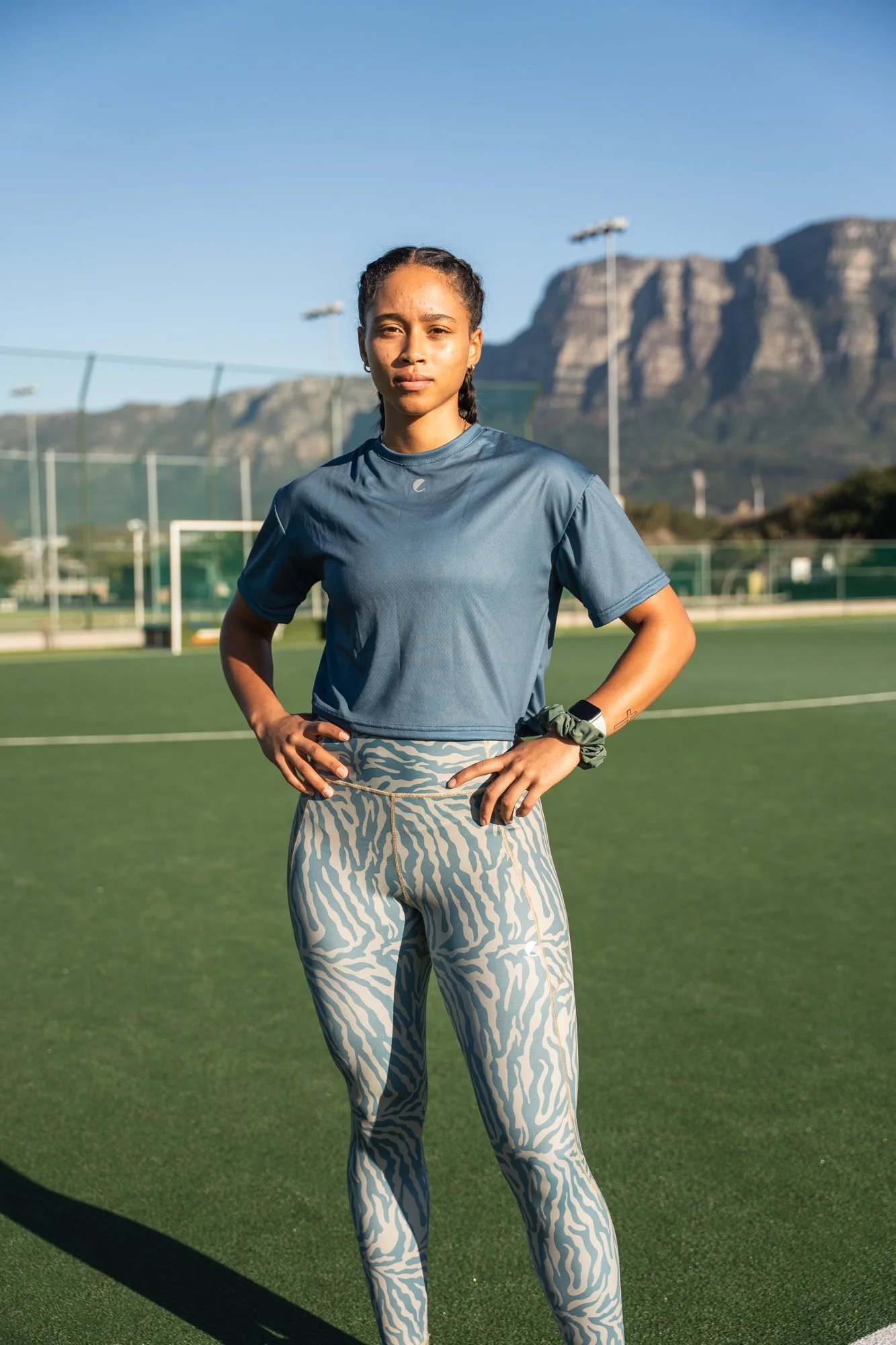 A young woman standing on a green sports field with her hands on her hips, wearing a blue athletic shirt and zebra-print pants, with a mountain backdrop and clear blue sky.