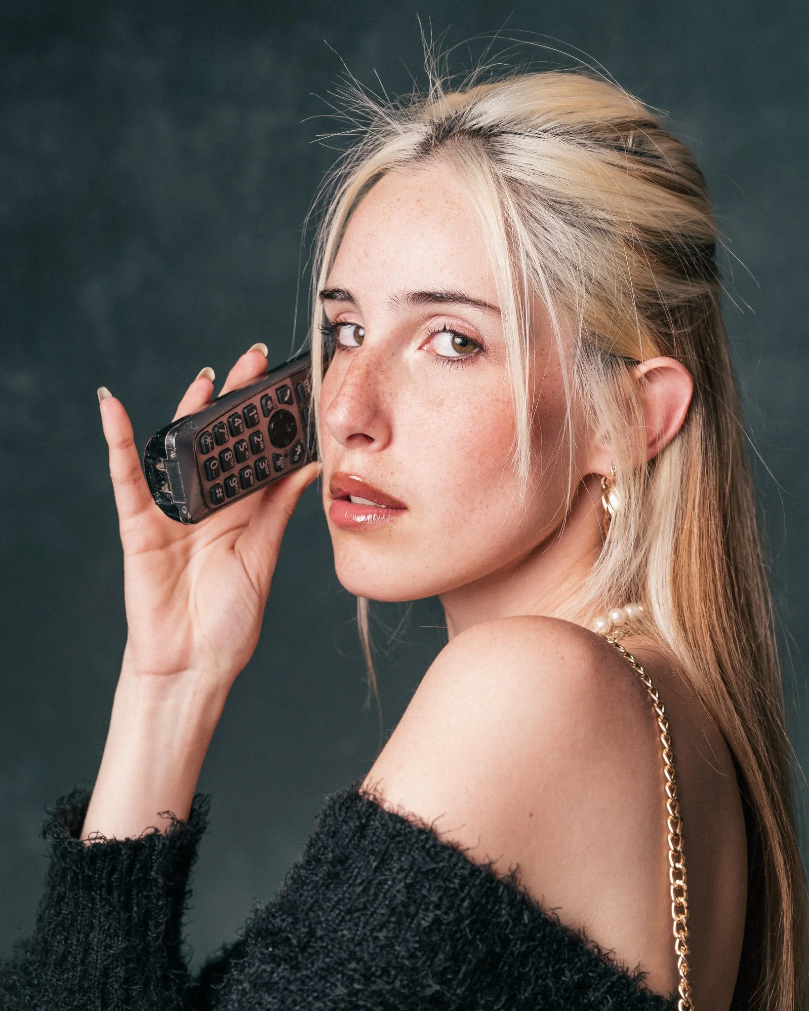 A young woman with blonde hair, freckles, and brown eyes holding a phone to her ear, wearing a black fuzzy sweater, gold chain, pearl earrings, and a tattoo on her arm, against a dark background.