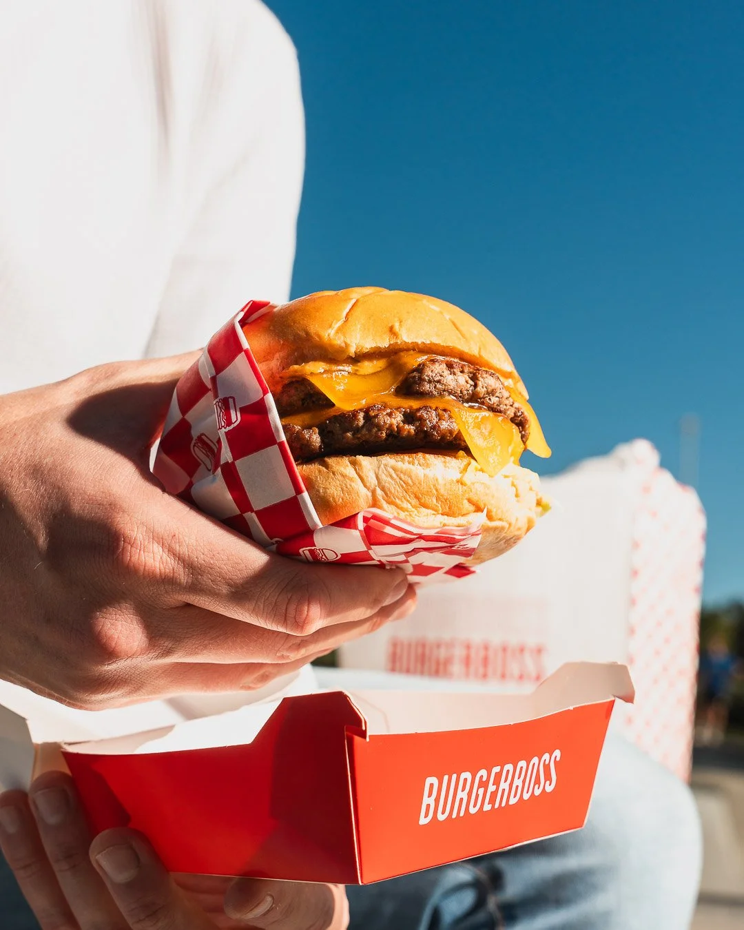 A person holding a cheeseburger in a red and white checkered wrapper with a blue sky in the background. The burger contains melted cheese, beef patties, and a sandwich bun. There is a BurgerBoss container visible at the bottom.