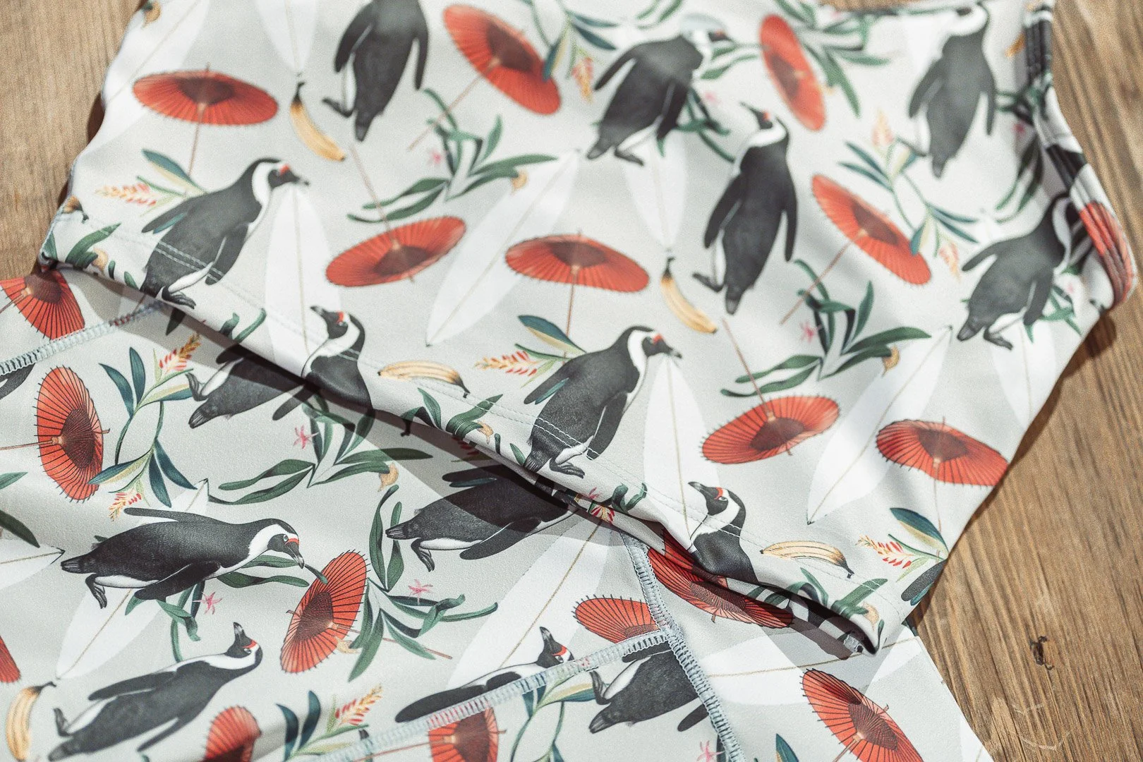 Close-up of a folded piece of clothing with a pattern of penguins holding umbrellas and bananas, lying on a wooden surface.