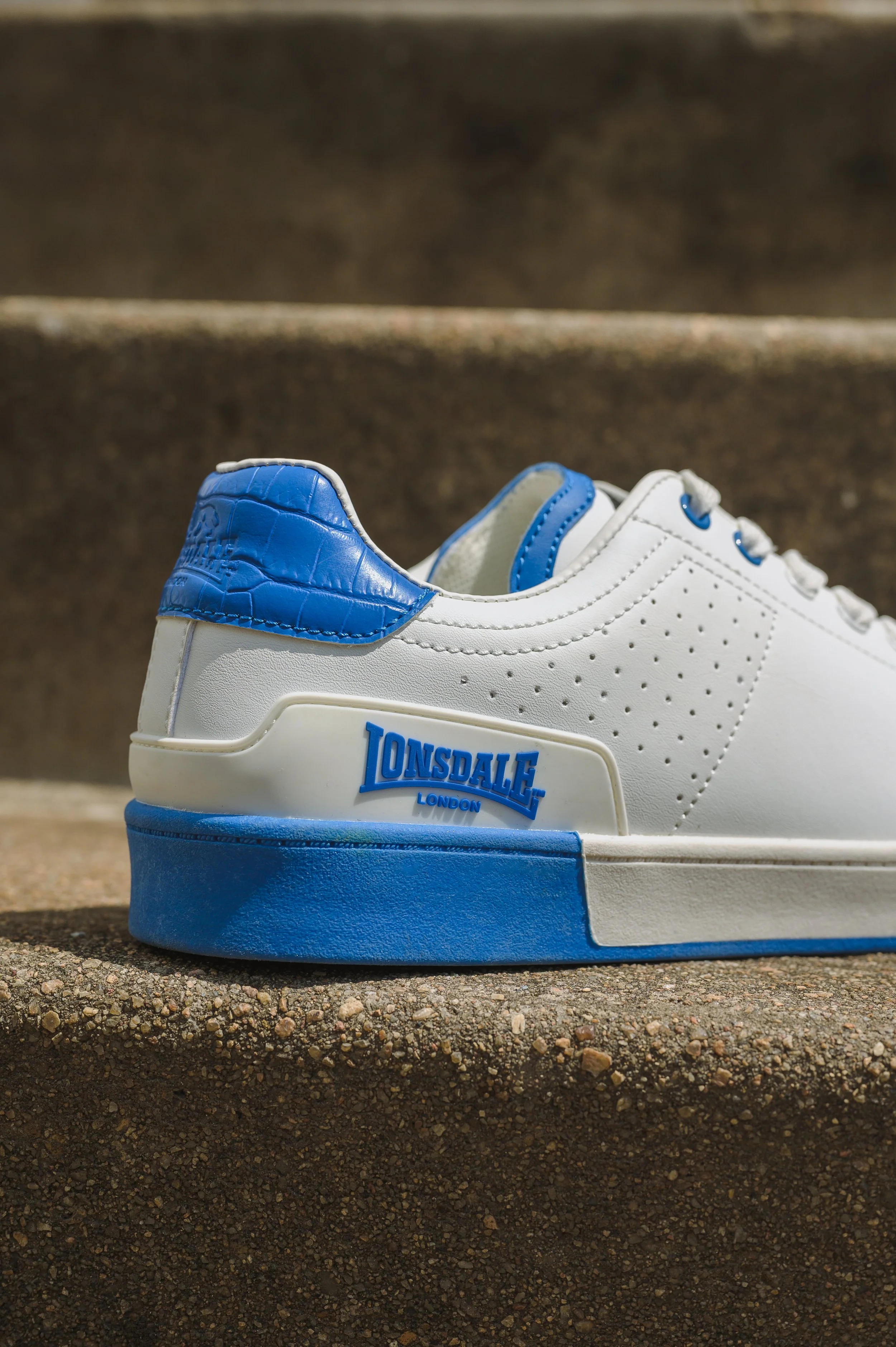 Close-up of a white Lonsdale London sneaker with blue accents on the heel and sole, placed on a textured concrete surface.