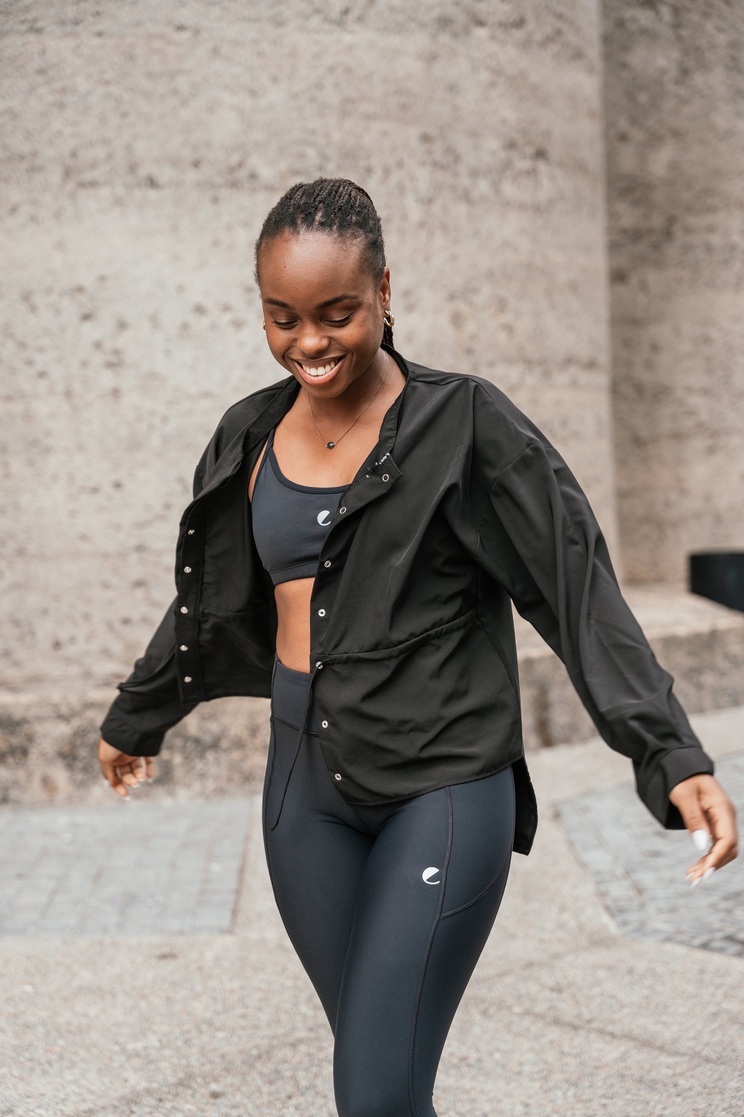 A woman smiling and looking down, wearing a black sports jacket and leggings, outdoors on a paved surface with a plain, beige wall in the background.