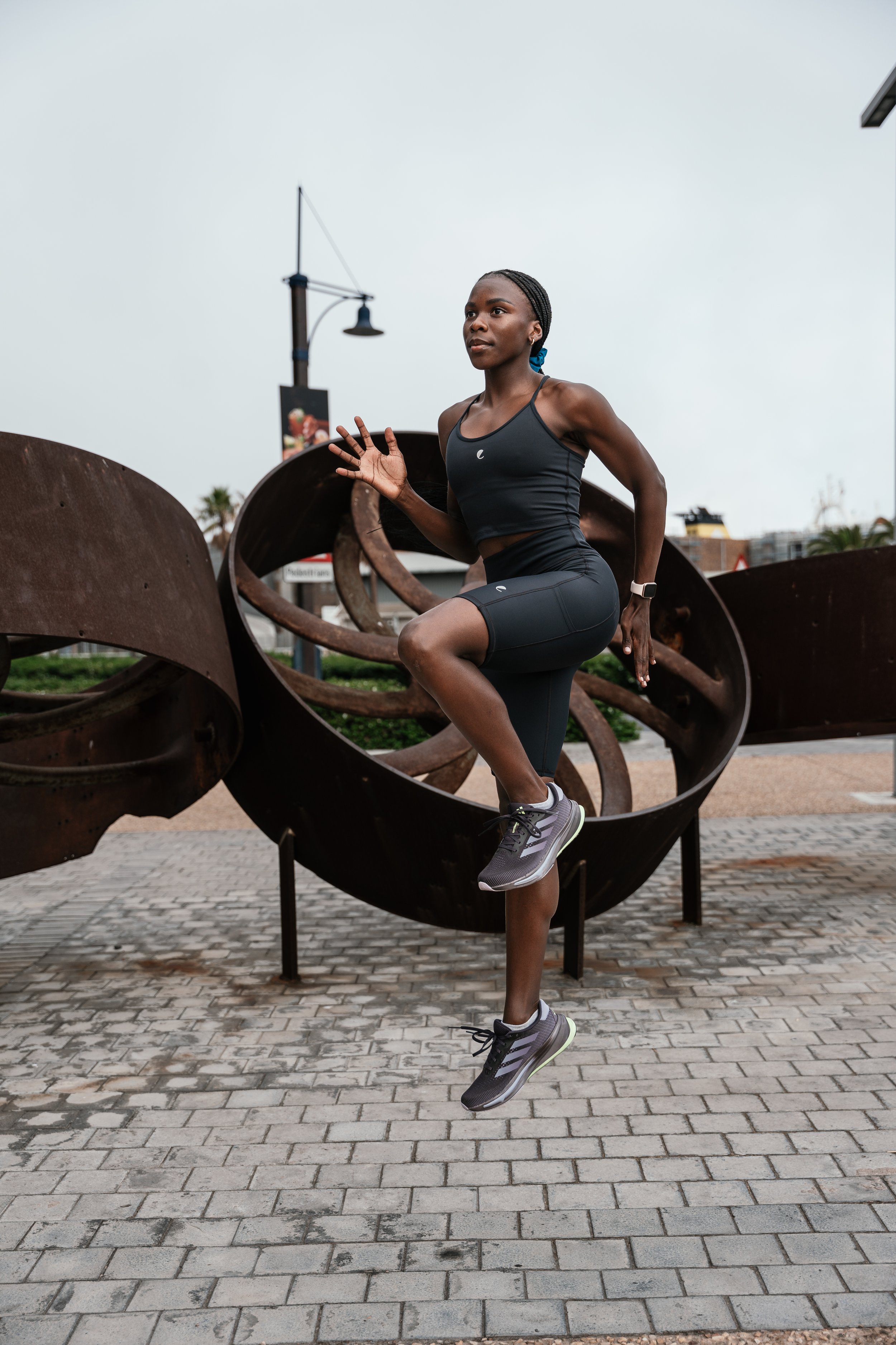 A woman in black workout attire, including leggings and a tank top, is running in an outdoor urban setting with sculptures and a lamppost in the background. She is wearing a smartwatch and running shoes, and has braided hair tied back with a blue ban