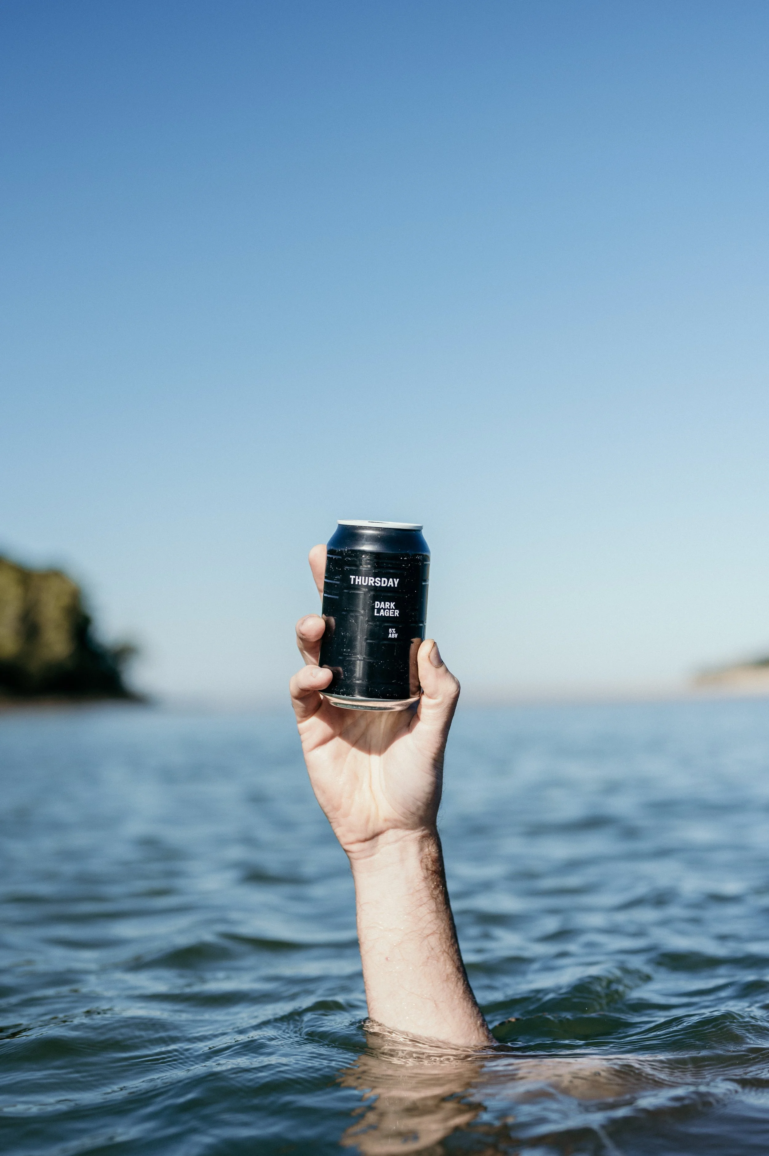 Person holding a black can labeled 'Thursday Dark Lager' above water, with a clear blue sky and distant shoreline in the background.