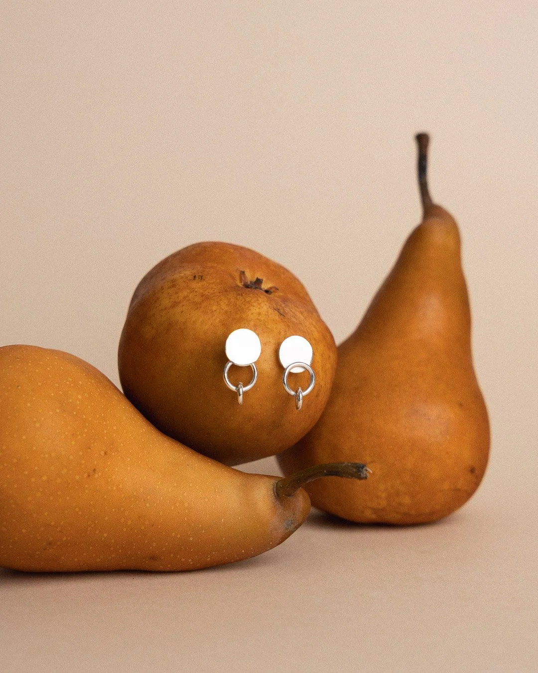A pair of silver hoop earrings with white circular posts, placed on a yellow pear and a red apple among other pears, on a beige background.