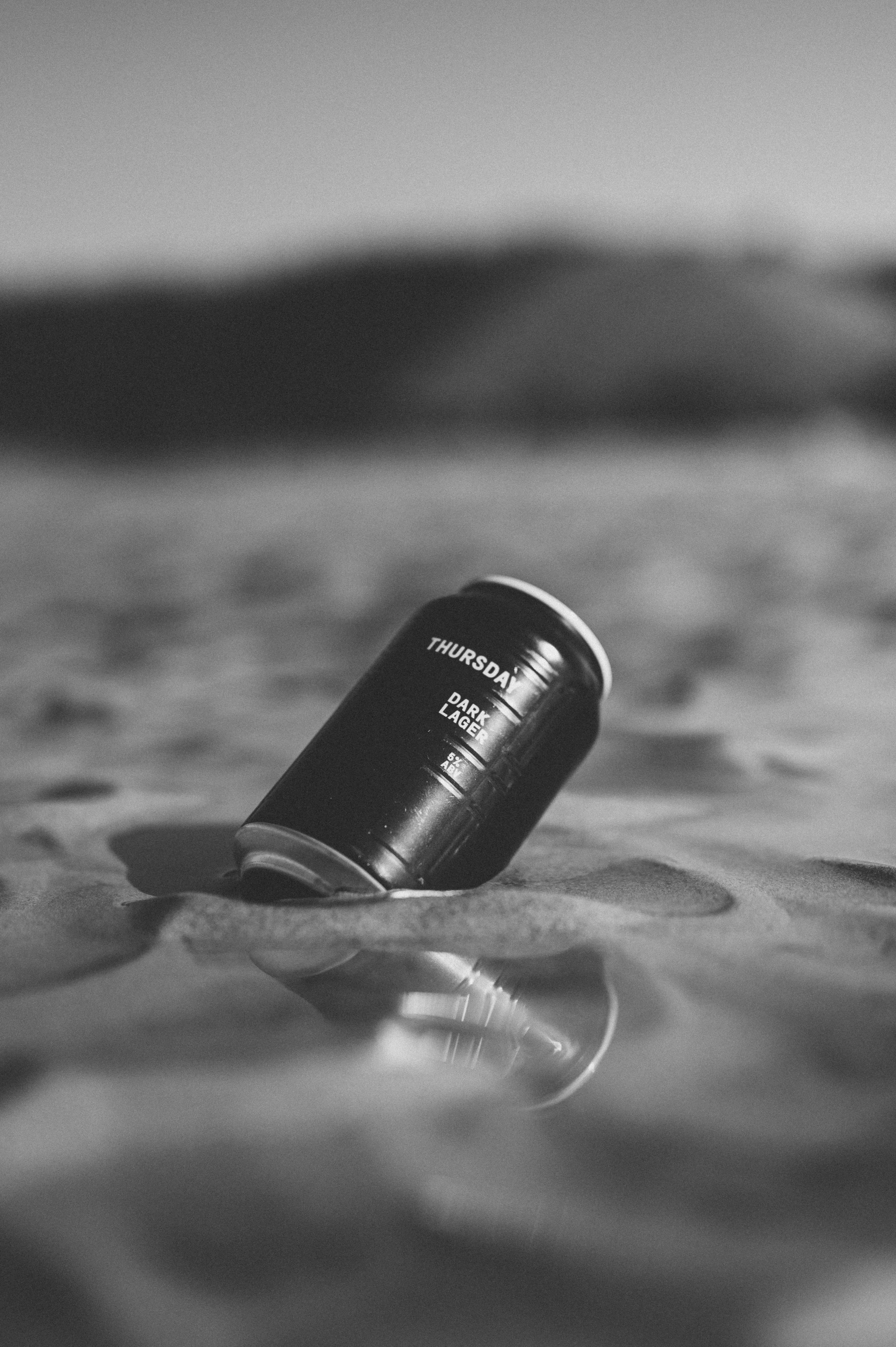 A black and white photo of a can of Thursday Dark Lager beer lying on its side on a sandy surface with a blurred background.
