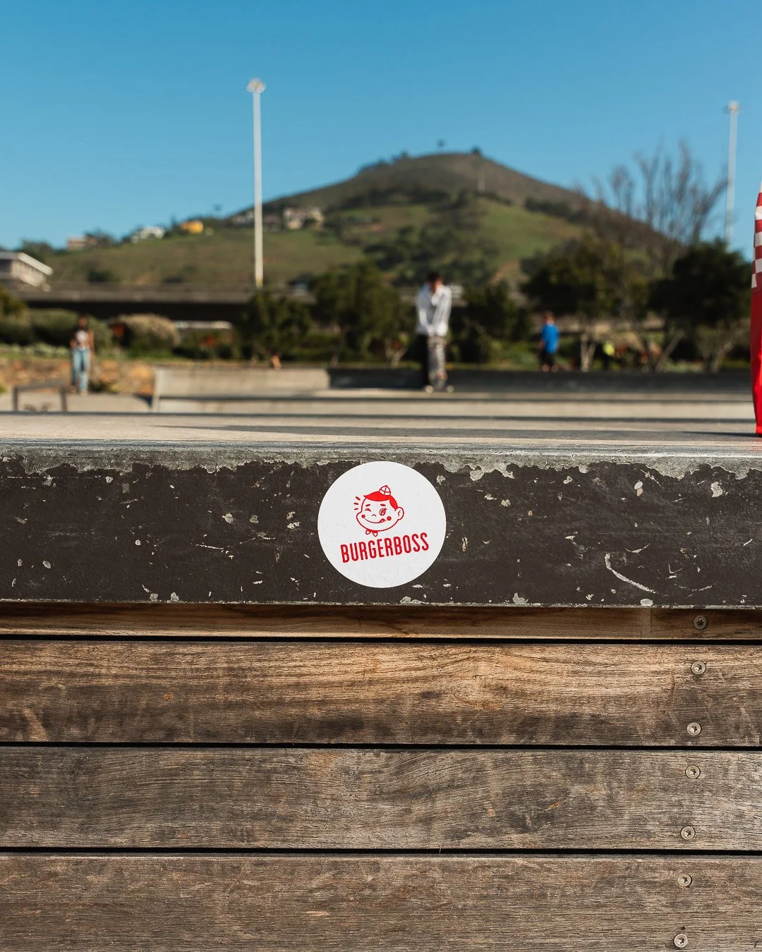 Close-up of a sticker that reads 'BURGERBOSS' with a cartoon face, on a wooden surface outdoors, with a park and hill in the background.